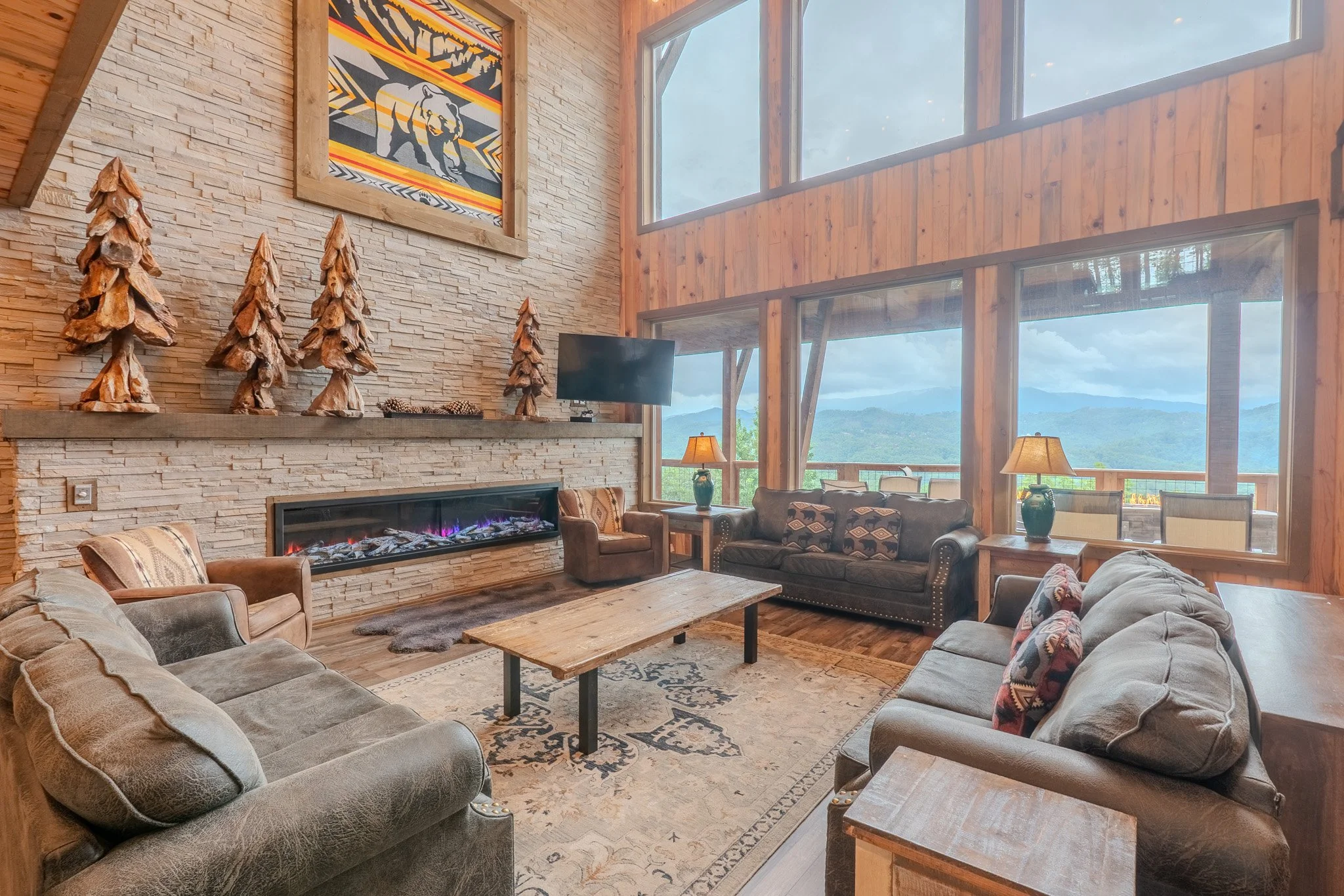 Living room with large windows showing mountain view, wood-paneled walls, stone fireplace, wooden sculptures of trees, leather and fabric sofas, wooden coffee table, lamps, and mountain-themed artwork.