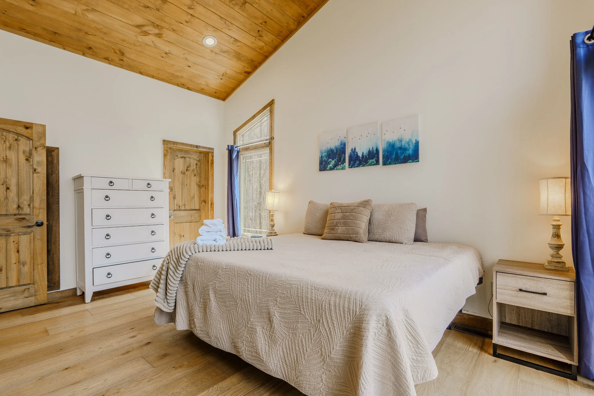 A cozy bedroom with a large bed covered in a cream quilt and multiple pillows, flanked by two nightstands with lamps, located next to a window with blue curtains. A white dresser is nearby, with folded towels on top, and the room features wood-panele