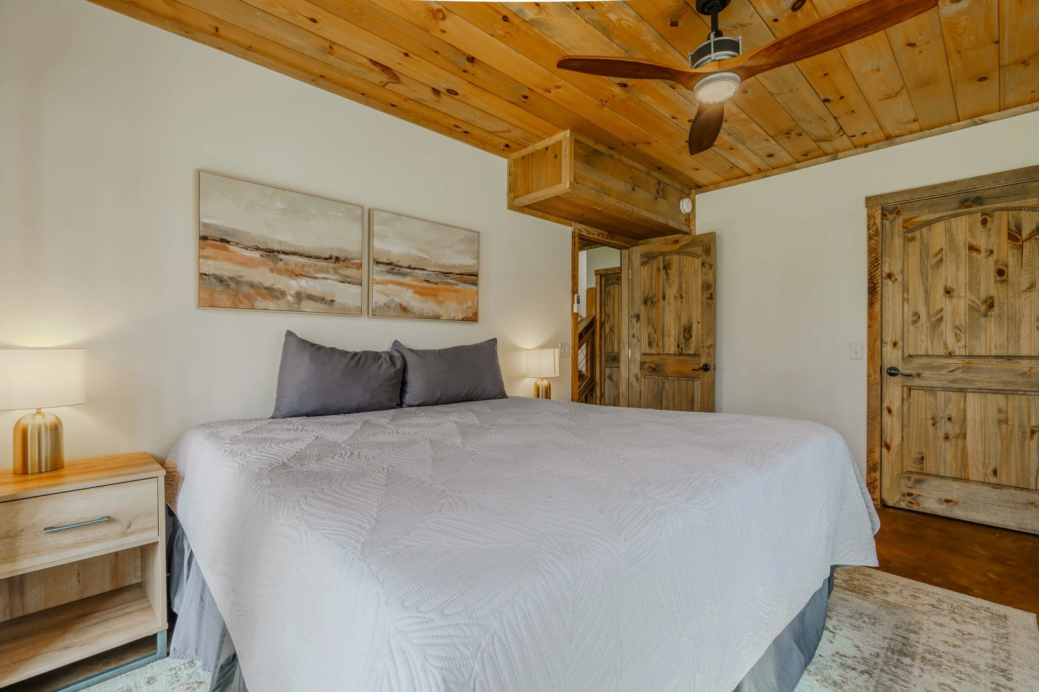 A bedroom with a wooden ceiling, two wooden doors, a large bed with gray pillows and white quilt, two bedside lamps, a painting above the bed, and a ceiling fan.