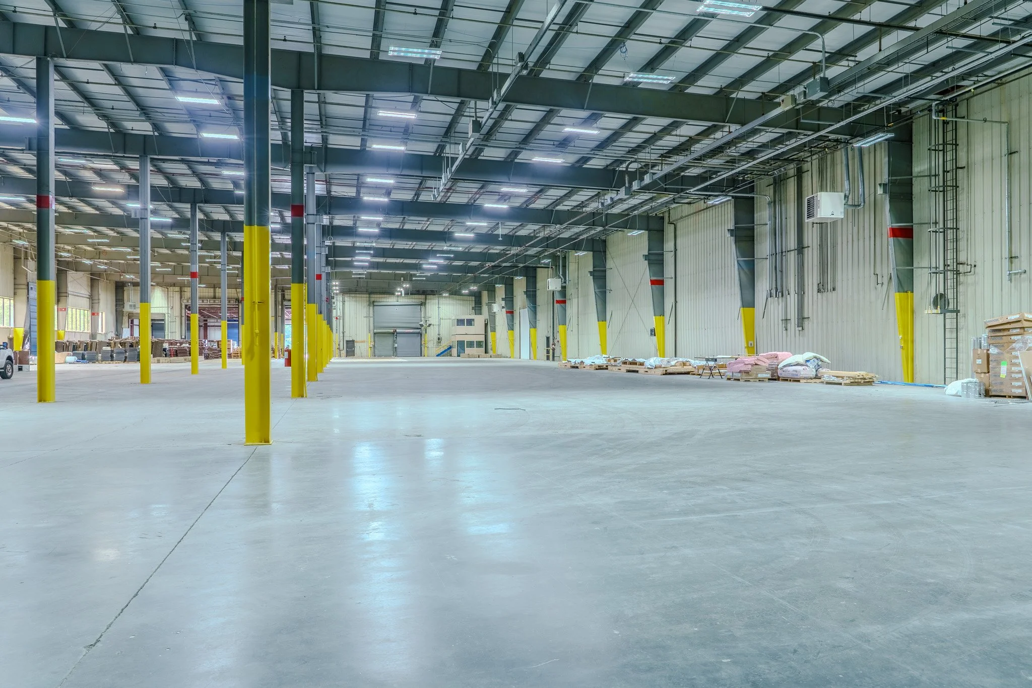 Empty warehouse with yellow safety posts, high ceiling, industrial lighting, and storage pallets along the walls.