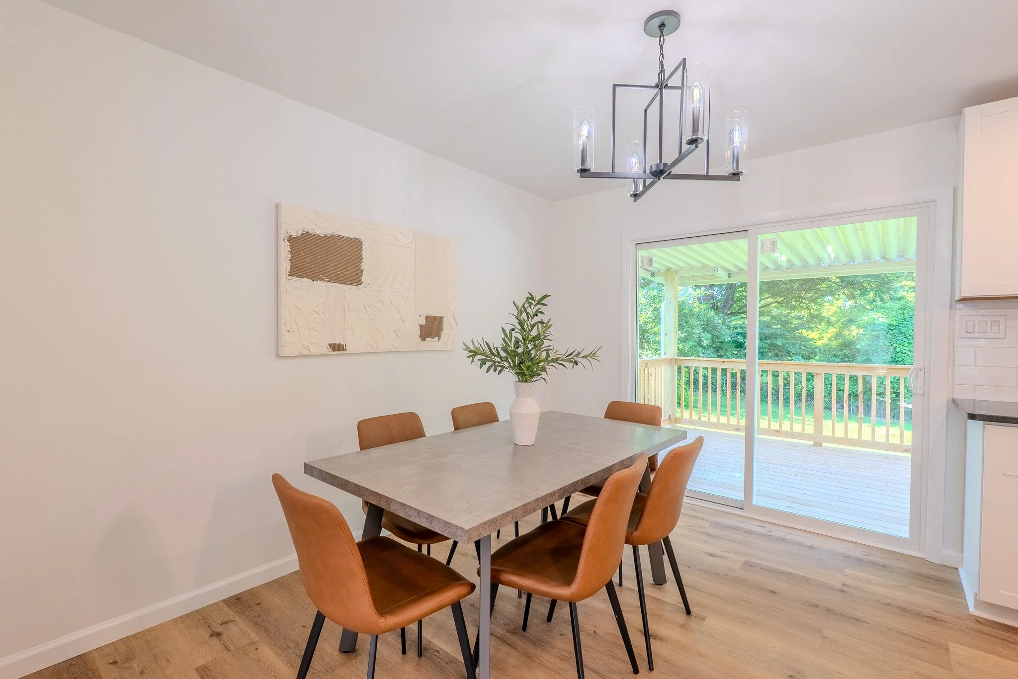 Dining room with a rectangular table, six brown chairs, a white vase with green branches on the table, a window showing a wooden deck and green trees outside, and a modern chandelier hanging from the ceiling.