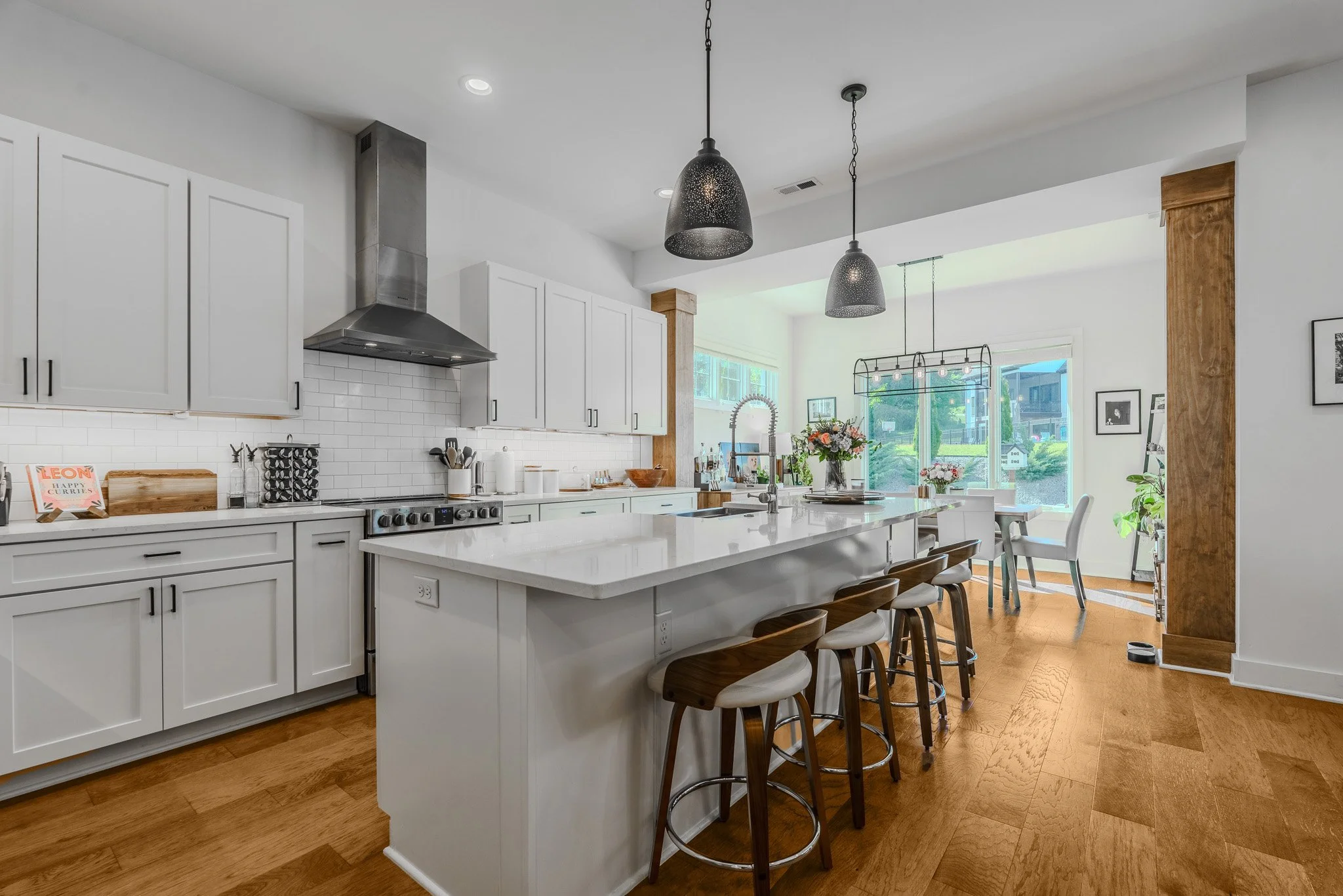 Bright, modern kitchen with white cabinets, a large island with bar stools, stainless steel appliances, and a dining area with large windows and outdoor views.
