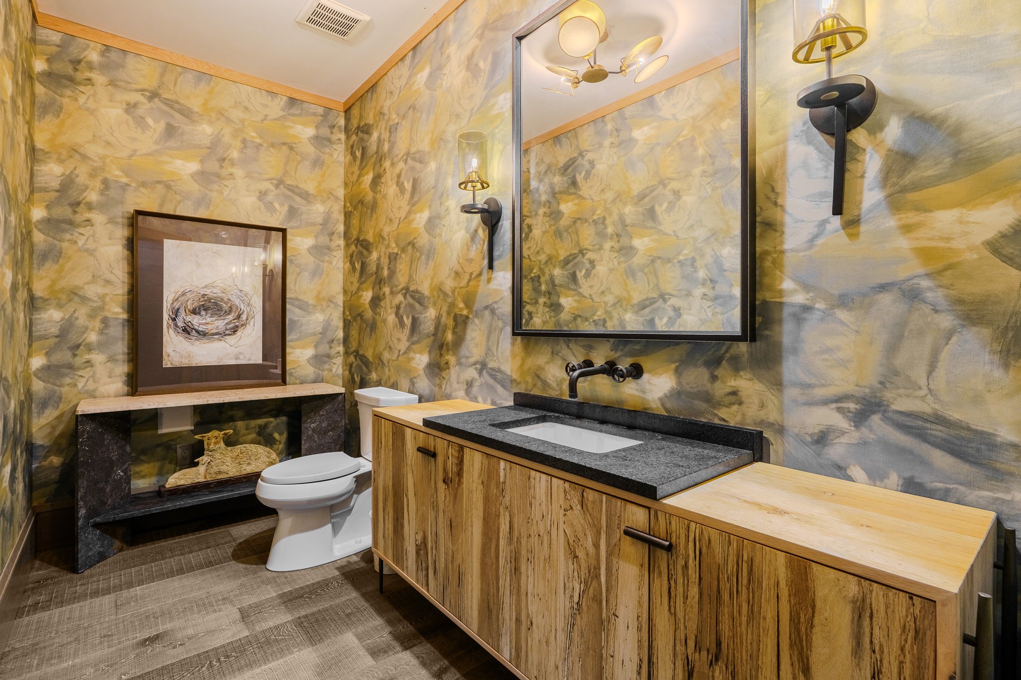 A modern bathroom with abstract yellow and gray painted walls, a large mirror, black wall sconces, a wooden vanity with a black countertop and white sink, a framed artwork, and wooden flooring.