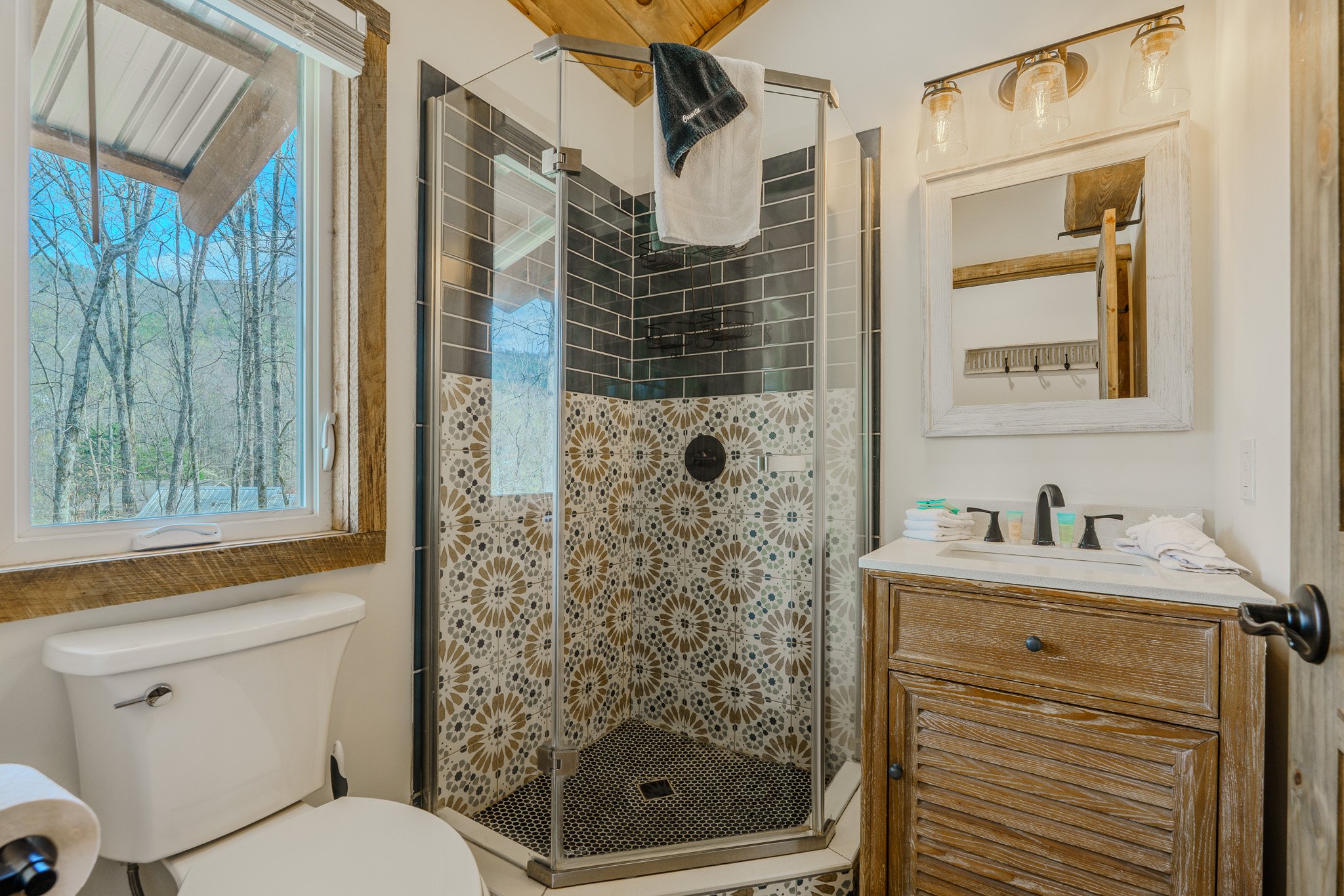 A small bathroom featuring a glass shower with patterned tile walls, a wooden vanity with a mirror, a toilet, and a window showing outdoor trees.