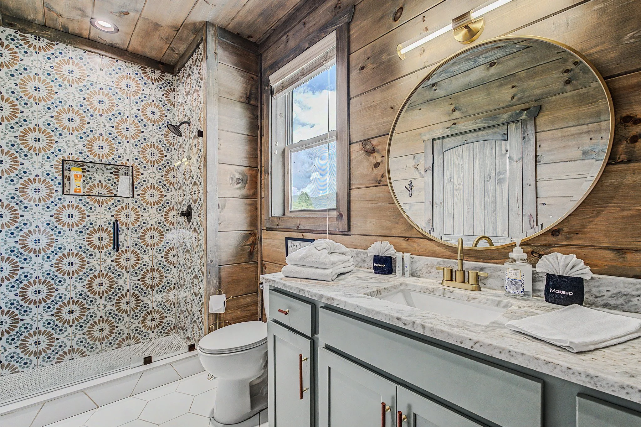 A rustic bathroom with wooden walls and ceiling, featuring a white marble countertop and large oval mirror. There are neatly folded towels, toiletries, and black aesthetic containers on the vanity. A window with a view of blue sky and clouds illumina