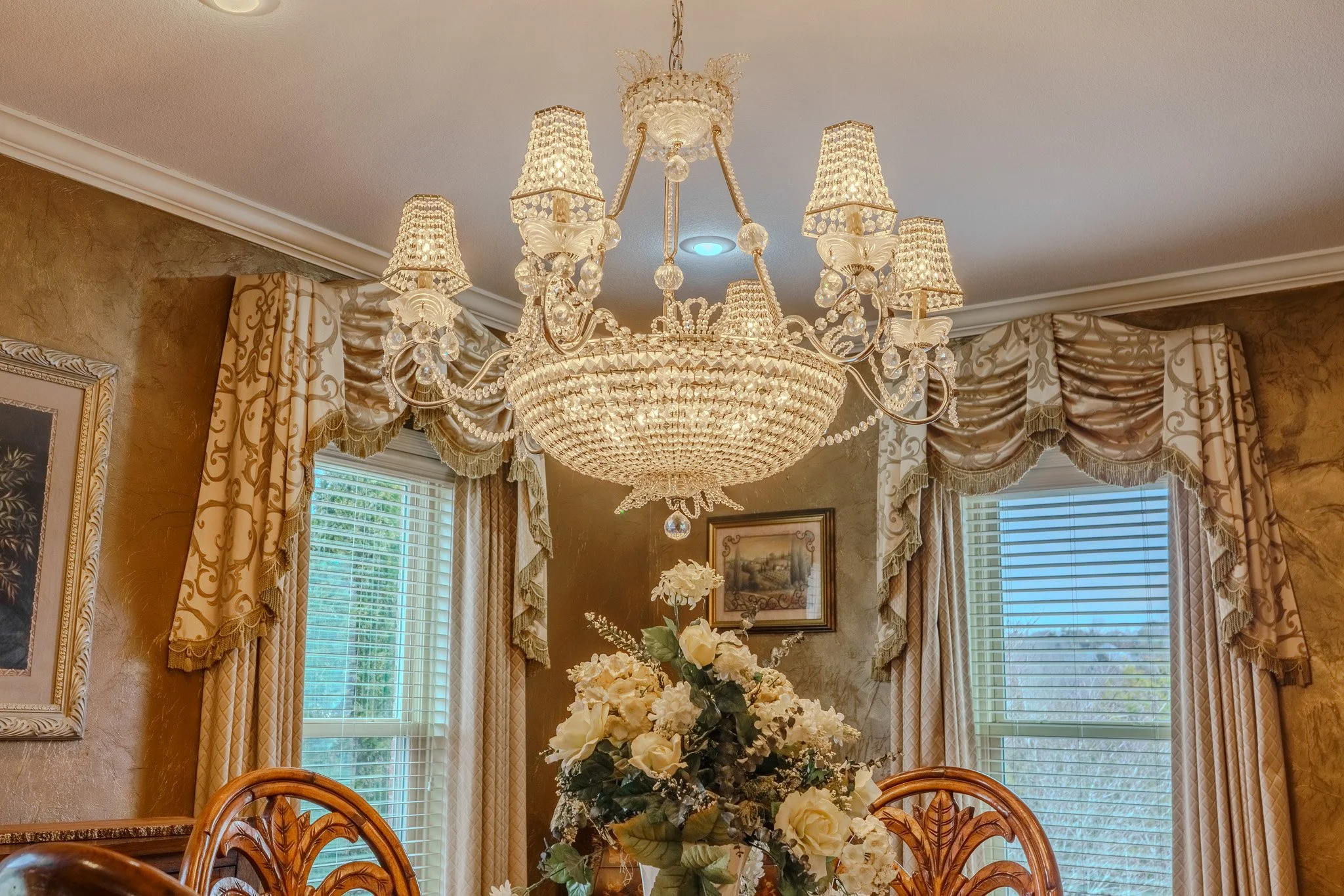 A luxurious dining room with a large chandelier hanging from the ceiling, floral curtains on windows, and a floral centerpiece on the table.