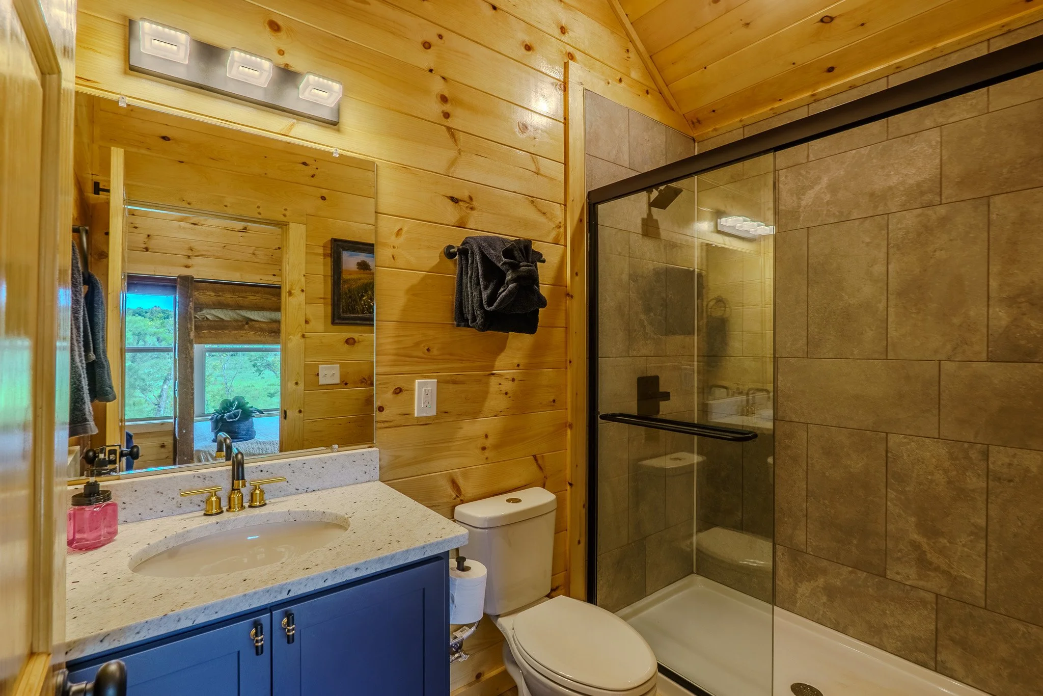 A rustic bathroom with wooden walls, a mirror above a light-colored countertop with a built-in sink, blue cabinet, toilet with a roll of toilet paper, a black towel hanging on the wall, and a glass-enclosed shower with brown tiles.