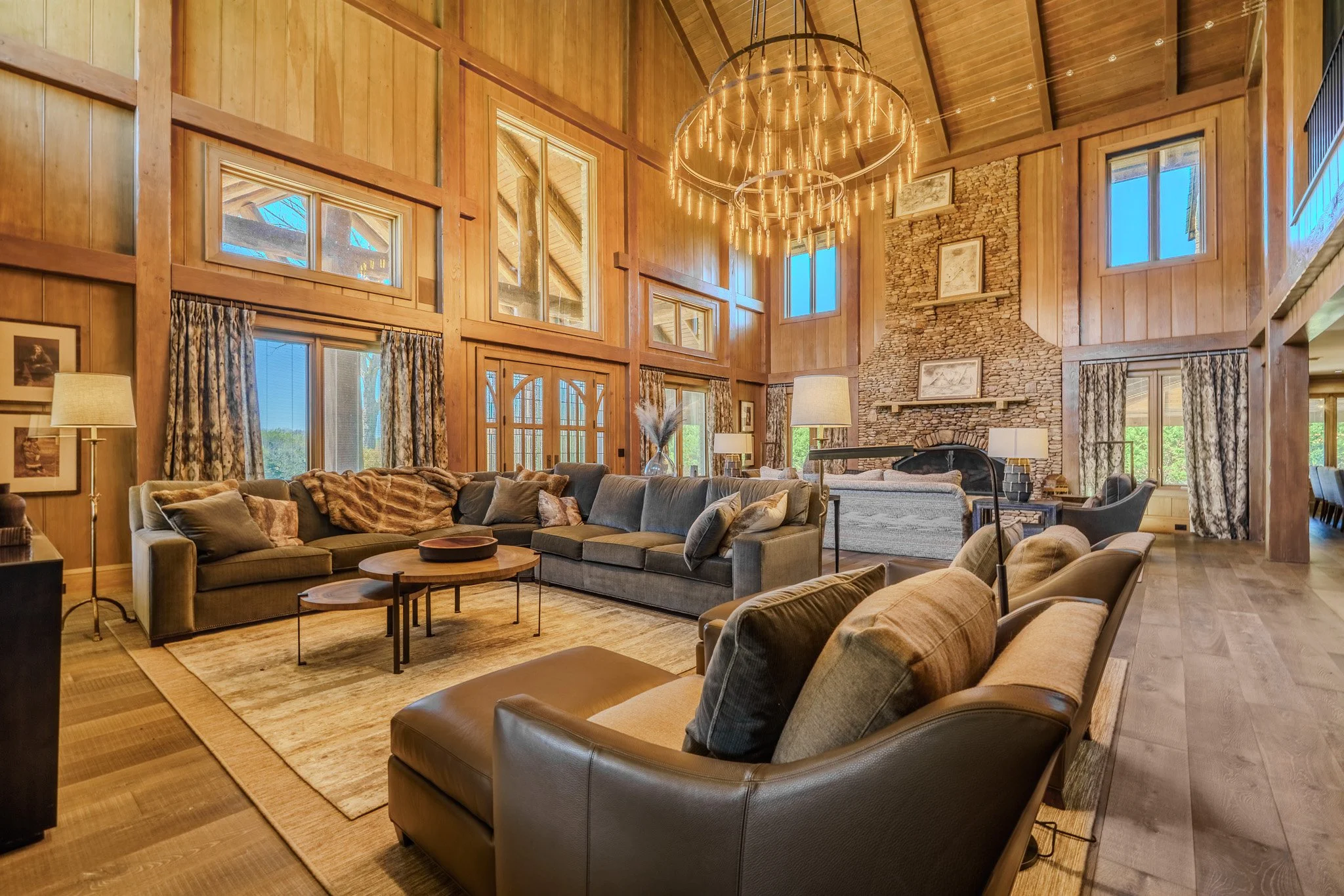 Spacious living room with wooden walls, large windows, a stone fireplace, and a high vaulted ceiling with a large circular chandelier.