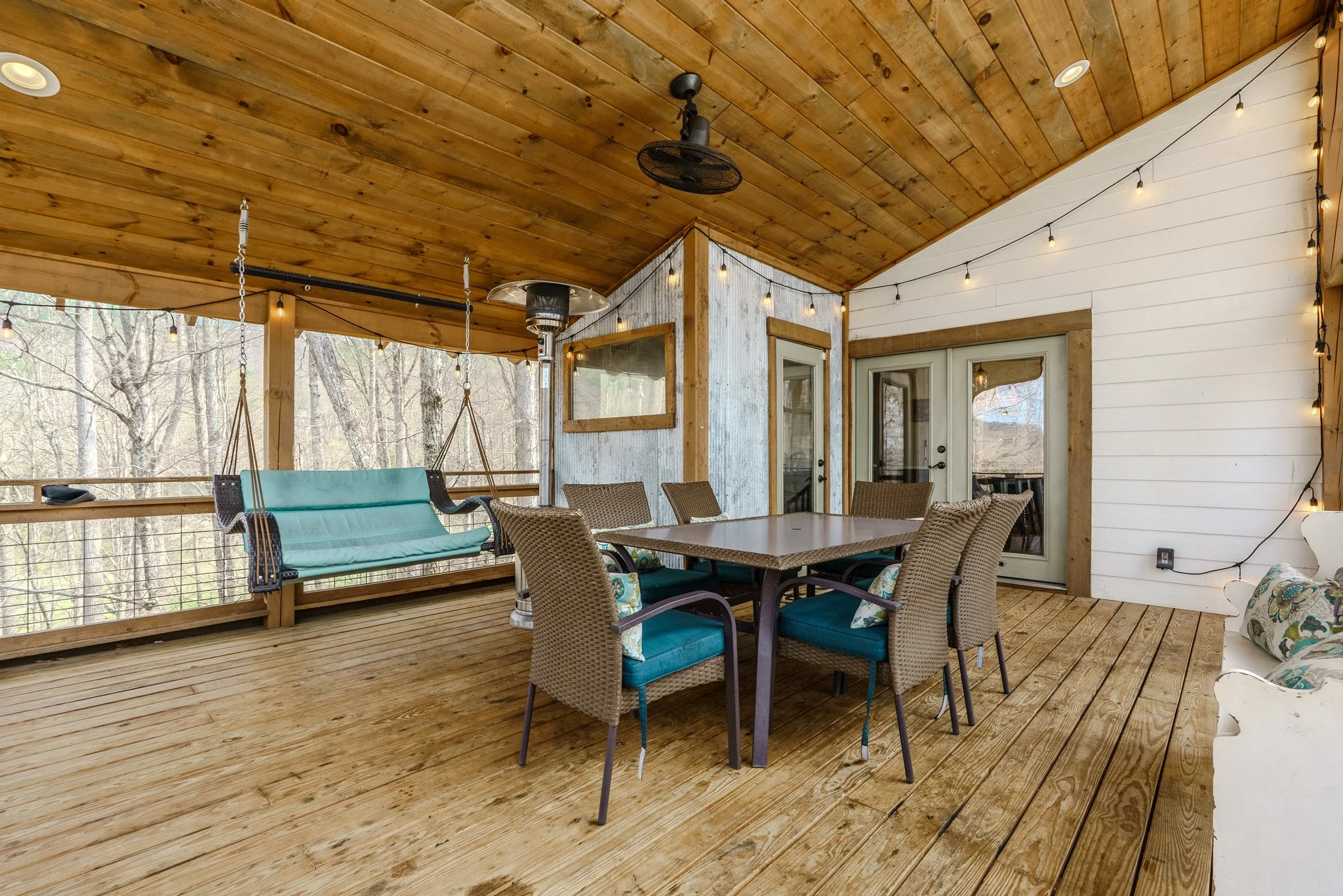 Covered wooden deck with outdoor dining table and six chairs, a porch swing, string lights, a ceiling fan, and a view of trees outside.