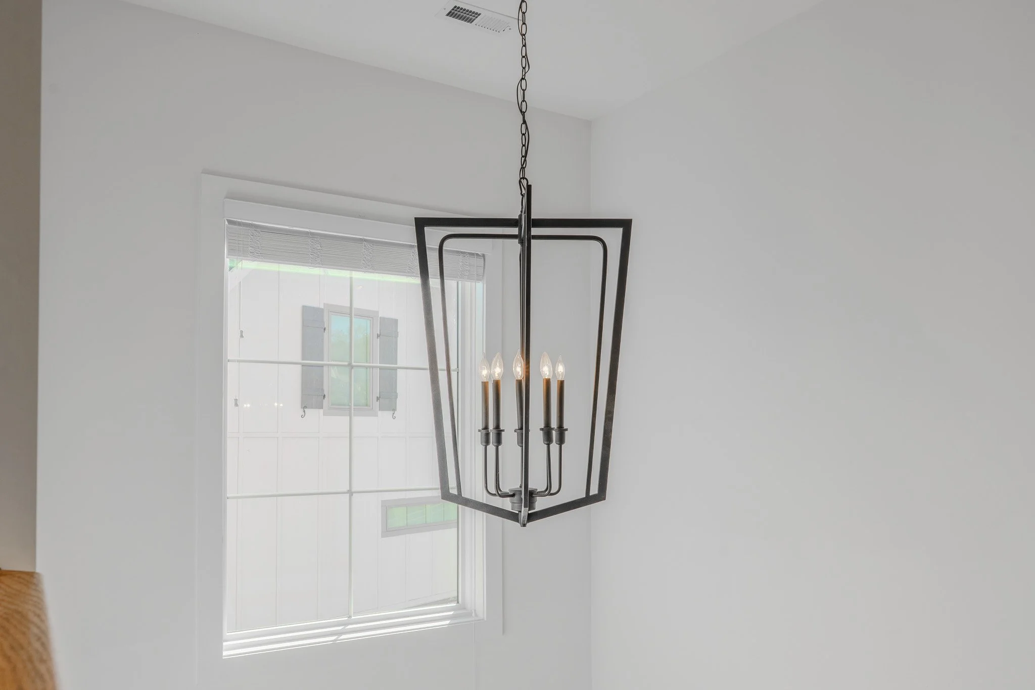 Modern black chandelier with five candles hanging from a chain in a white room with a large window.