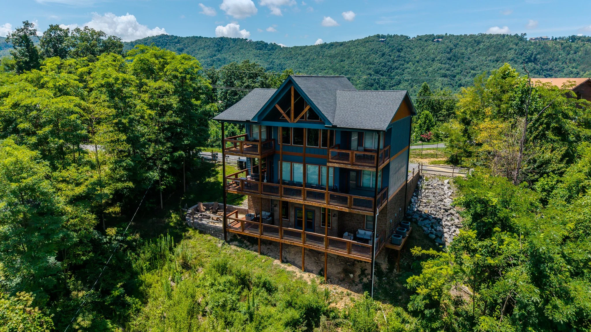 A multi-story house with blue and yellow exterior, surrounded by lush green trees and hills, featuring a large wooden deck on each level.