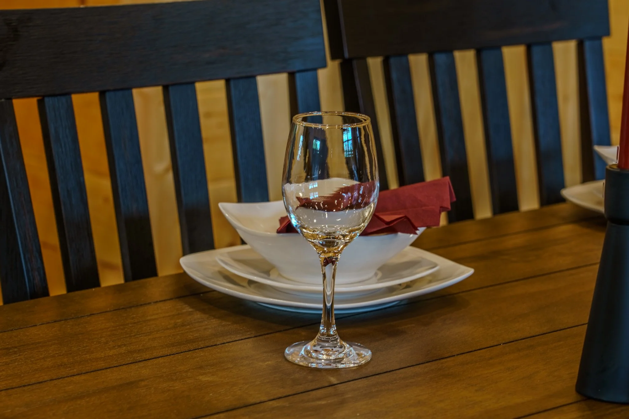 An empty wine glass on a wooden table, with a white bowl, a red napkin, and plates in the background.