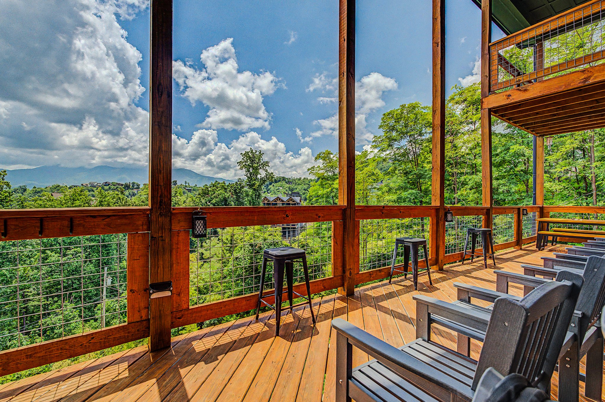 Balcony with wooden flooring and railing overlooking lush green trees and distant mountains under a partly cloudy sky.