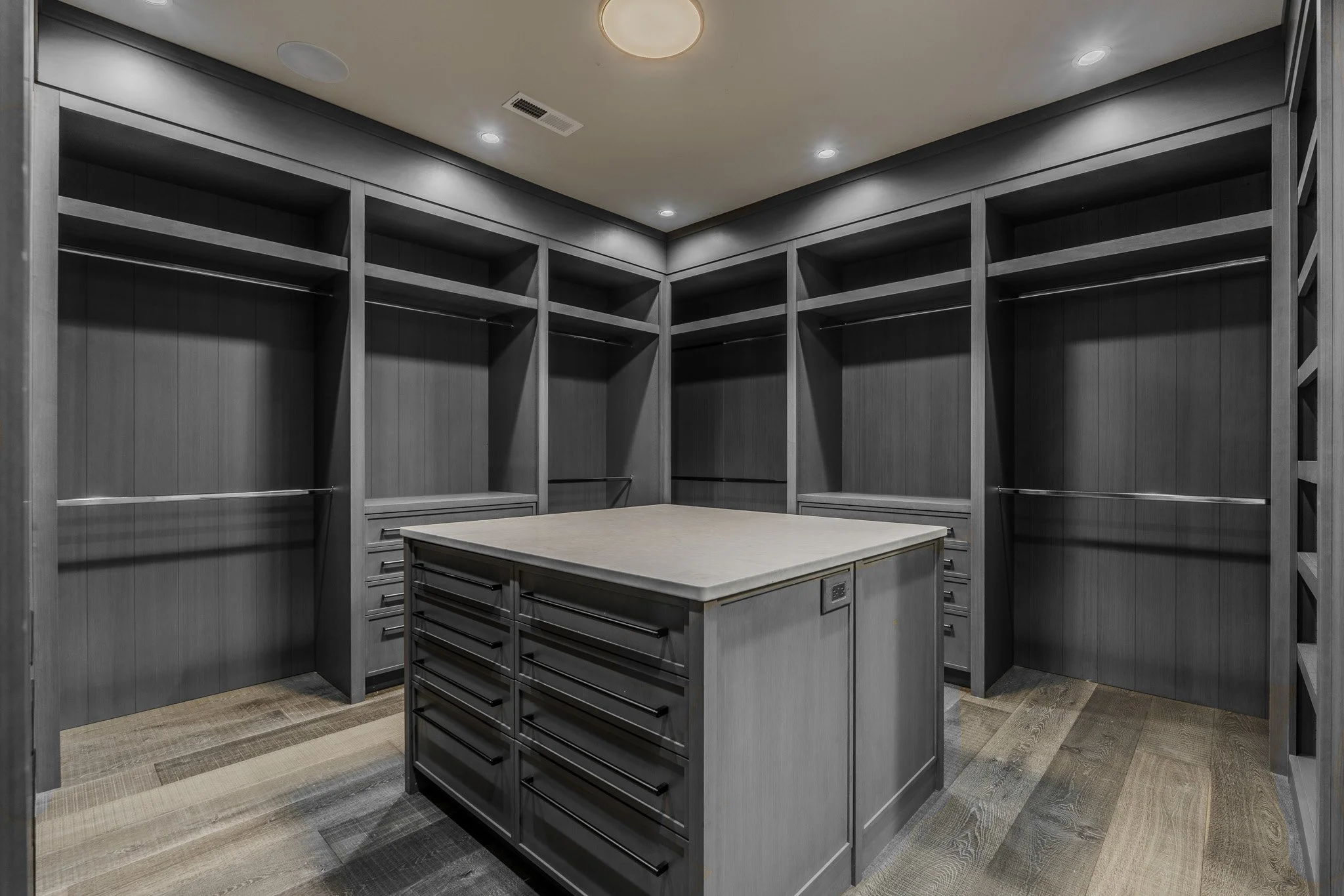 Empty walk-in closet with dark gray wood shelves, drawers, and an island with multiple drawers, hardwood flooring, and recessed ceiling lights.