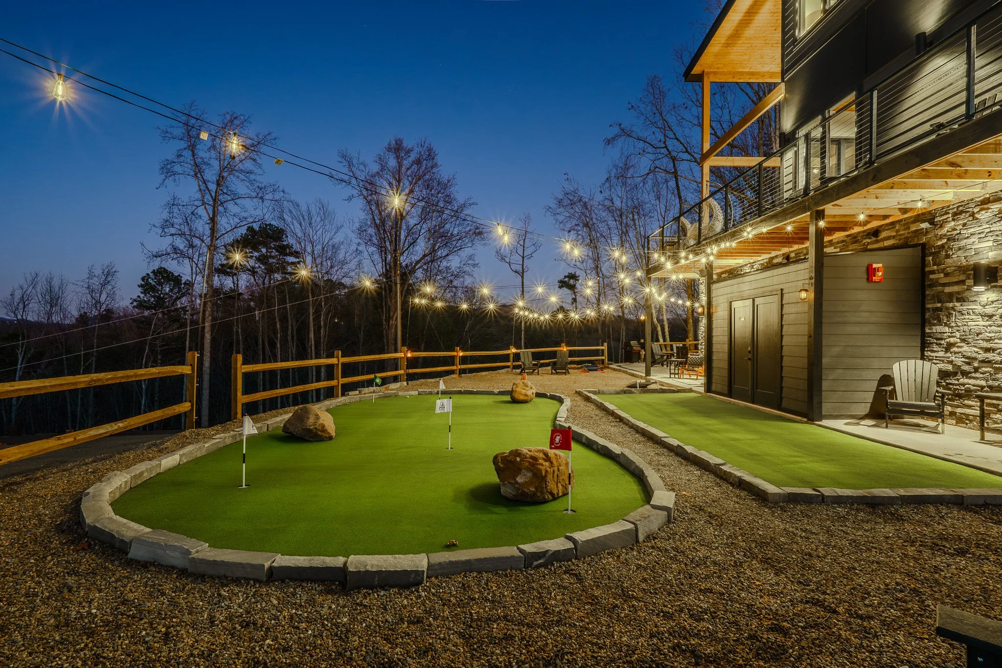 A mini-golf course at dusk with string lights, rocks, and flag holes, adjacent to a modern house with wood and stone exterior.