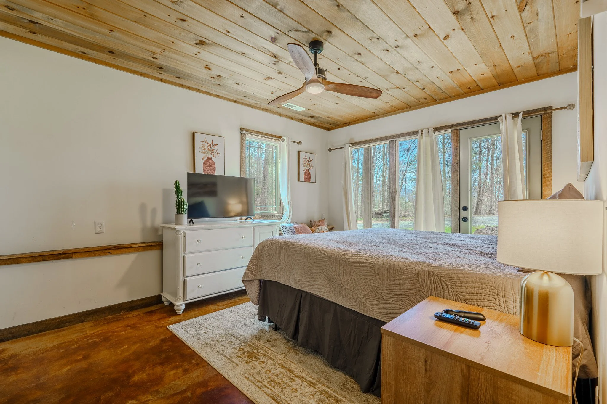 A bedroom with a wooden ceiling and floor, a large bed with a beige quilt and two remote controls on the nightstand, a white dresser with a TV and potted cactus, two framed pictures of vases, and large windows with white curtains letting in natural l
