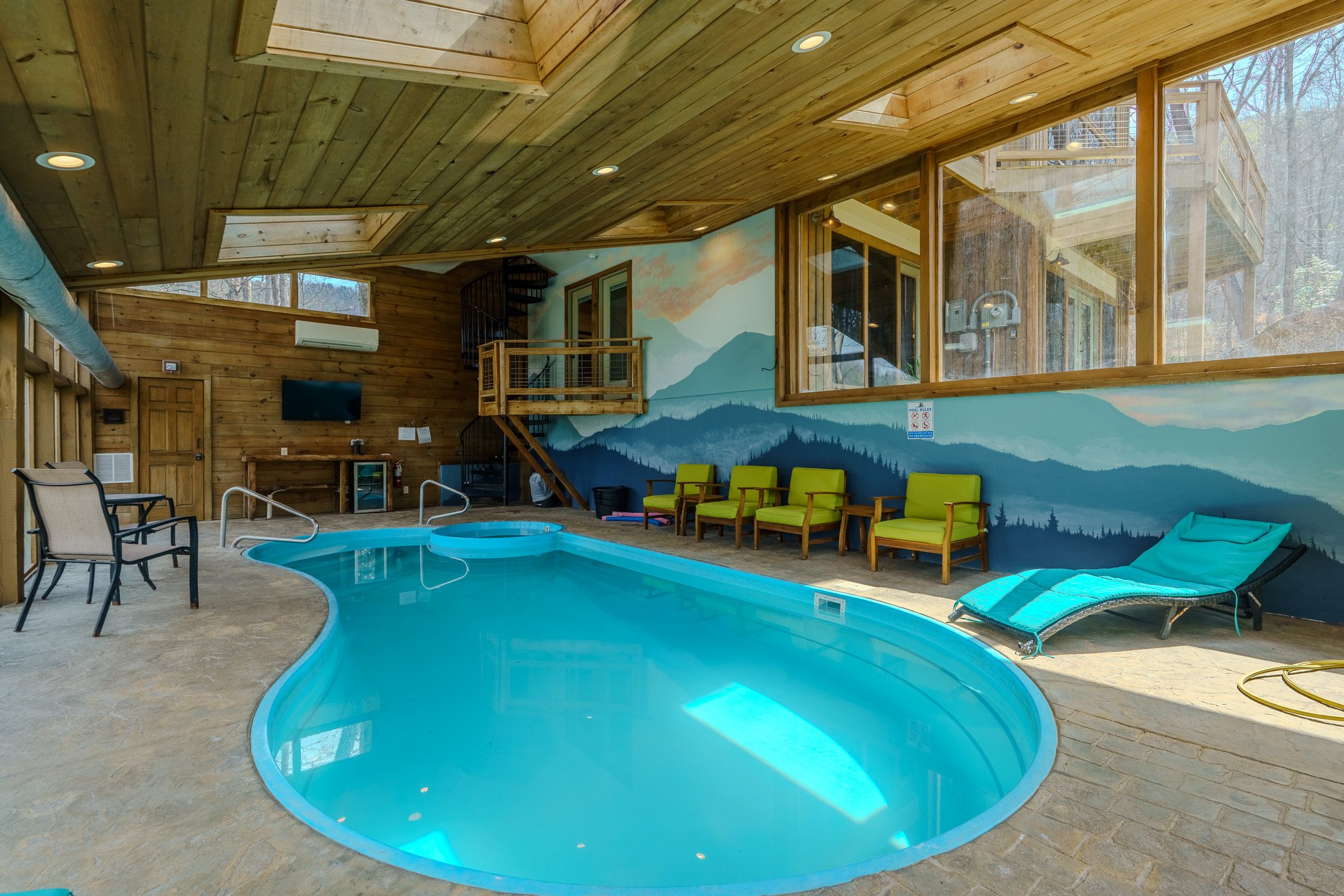 Indoor swimming pool area with wooden walls and ceiling, green chairs along one wall, lounge chair, a large window with mountain landscape mural, and a staircase leading to an upper level.