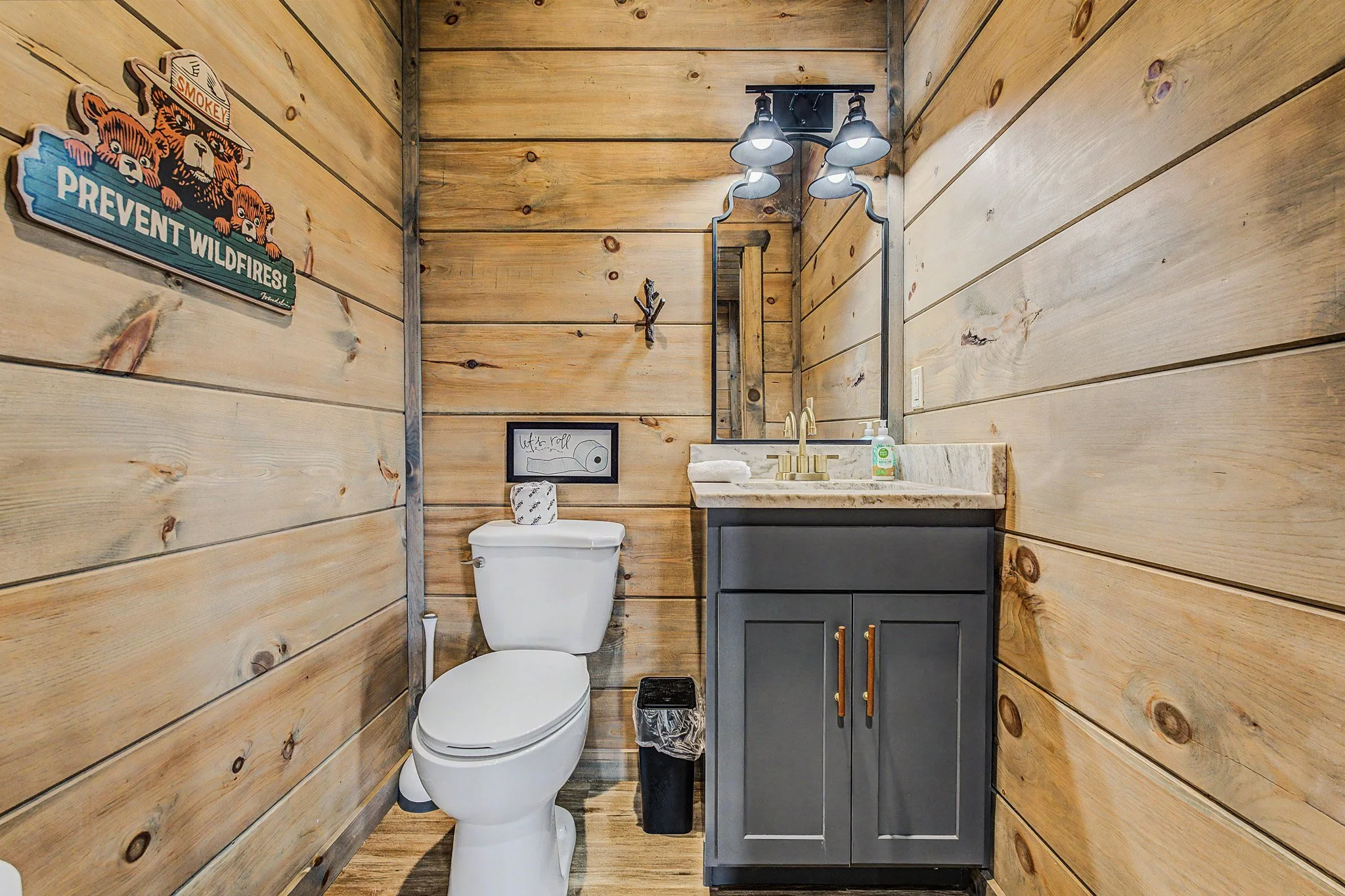 A small bathroom with wood paneling walls, a modern toilet, a dark gray vanity with a marble countertop, a mirror, a wall-mounted light fixture, and decorative wall art.