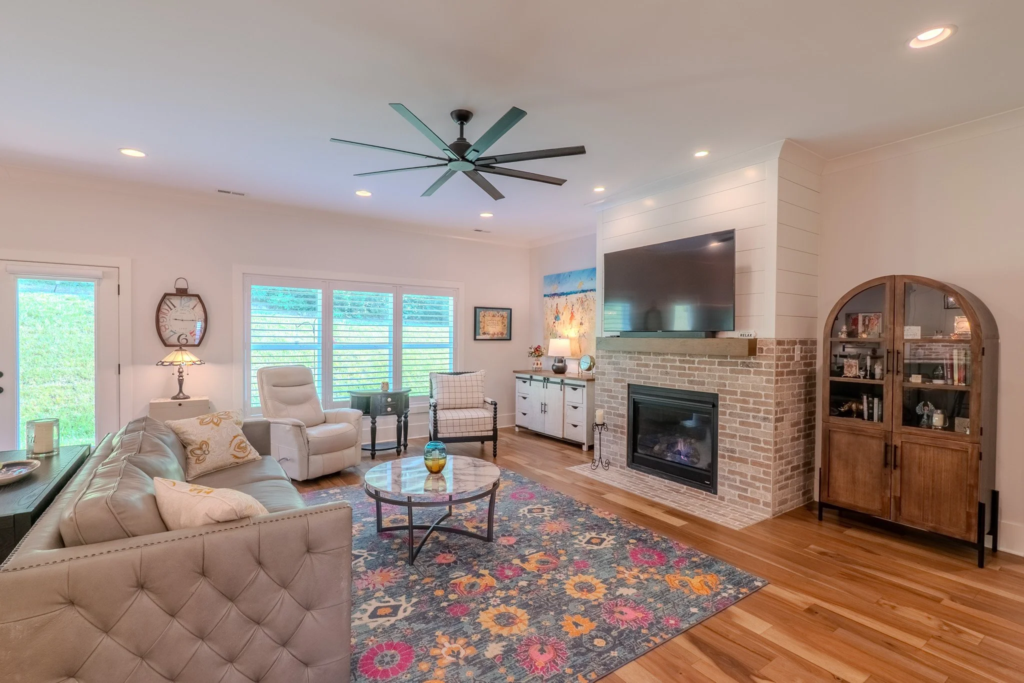 Living room with beige sofa, white armchair, wooden cabinet, brick fireplace, mounted TV, colorful rug, and large windows