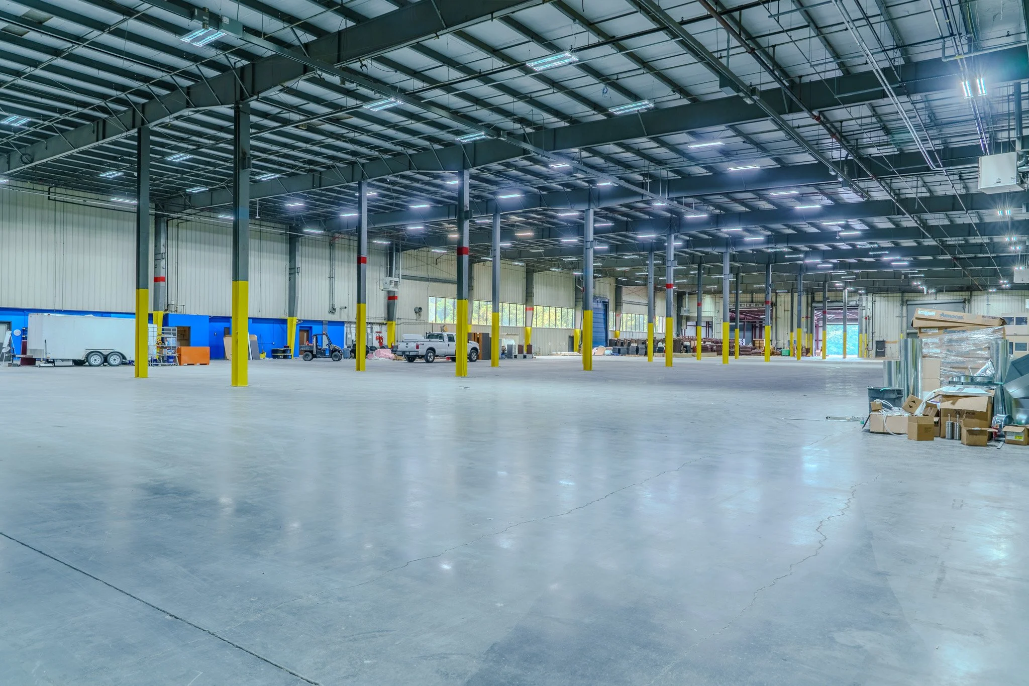 Large empty warehouse with high ceiling, concrete floor, vending machines, and several parked trucks along the wall.