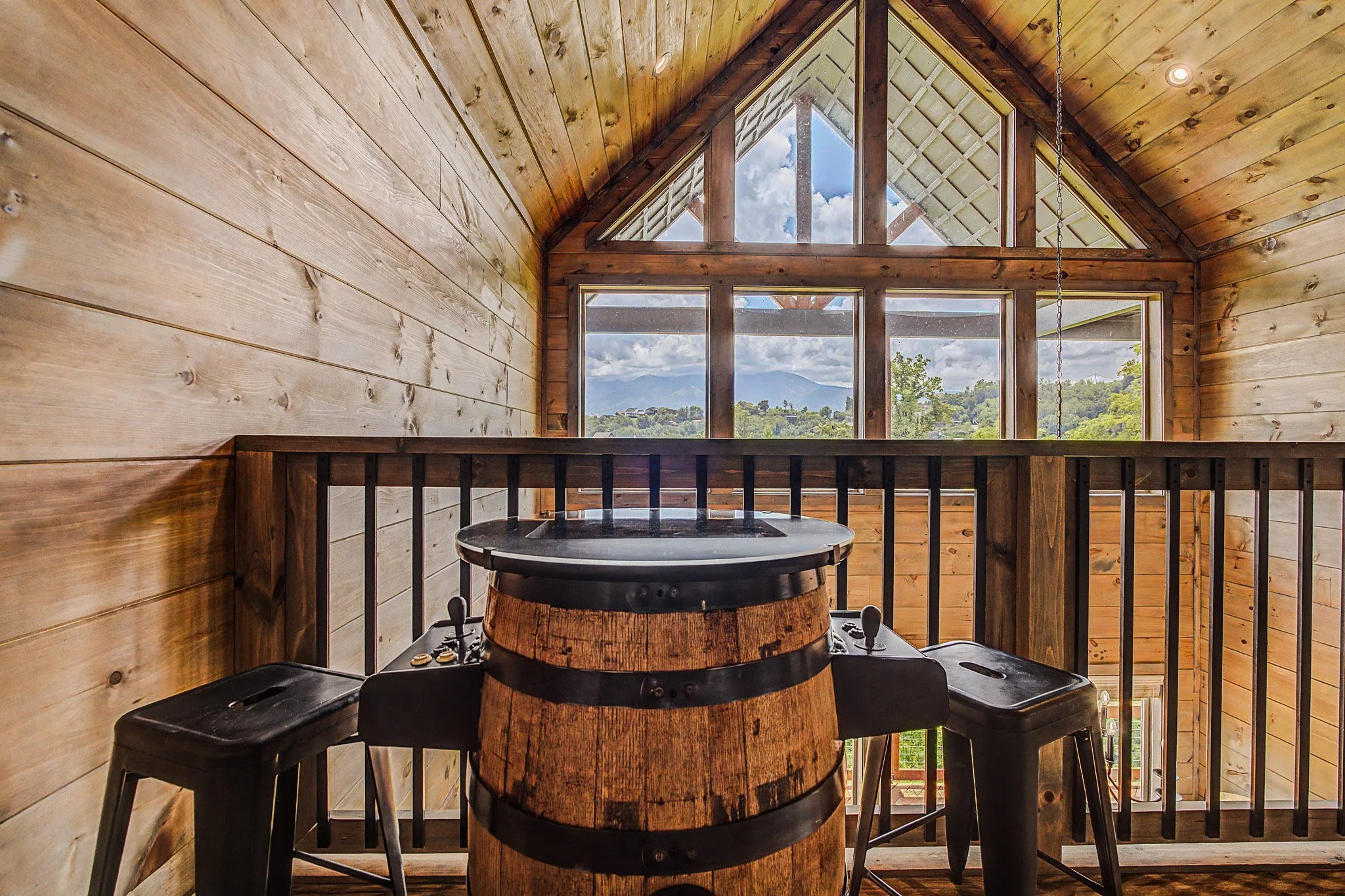 A cozy cabin interior with wooden walls and ceiling, large windows showing a mountain and cloudy sky, a DJ turntable on a wooden barrel, and three black chairs.
