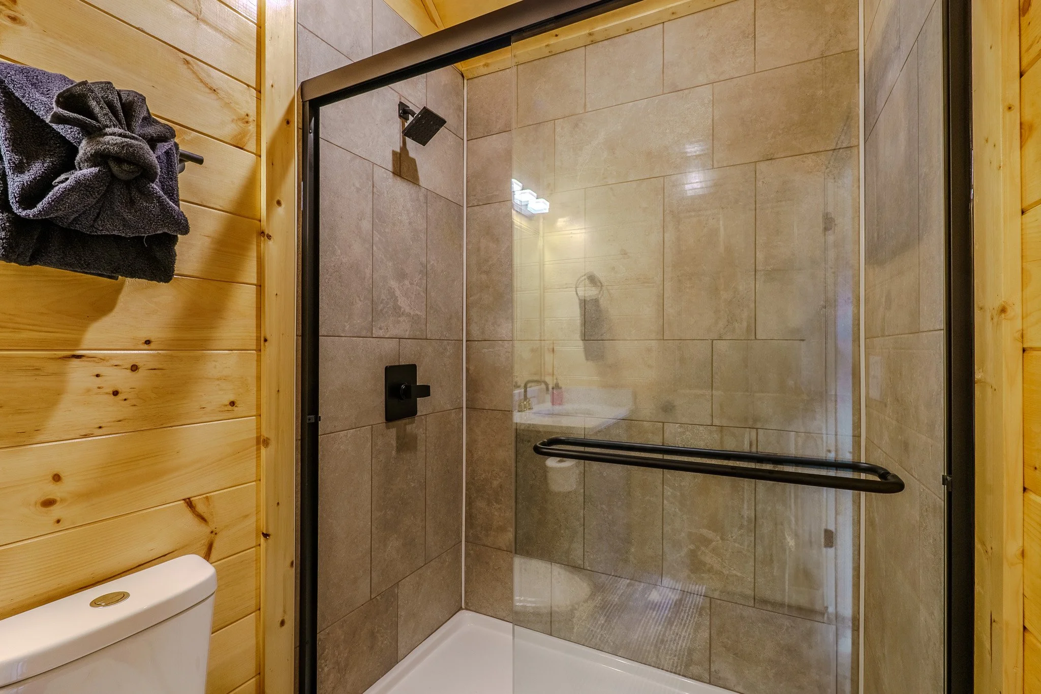 A walk-in shower with tan tiles, a glass door with a black handle and hardware, and a black showerhead on a wooden wall background.