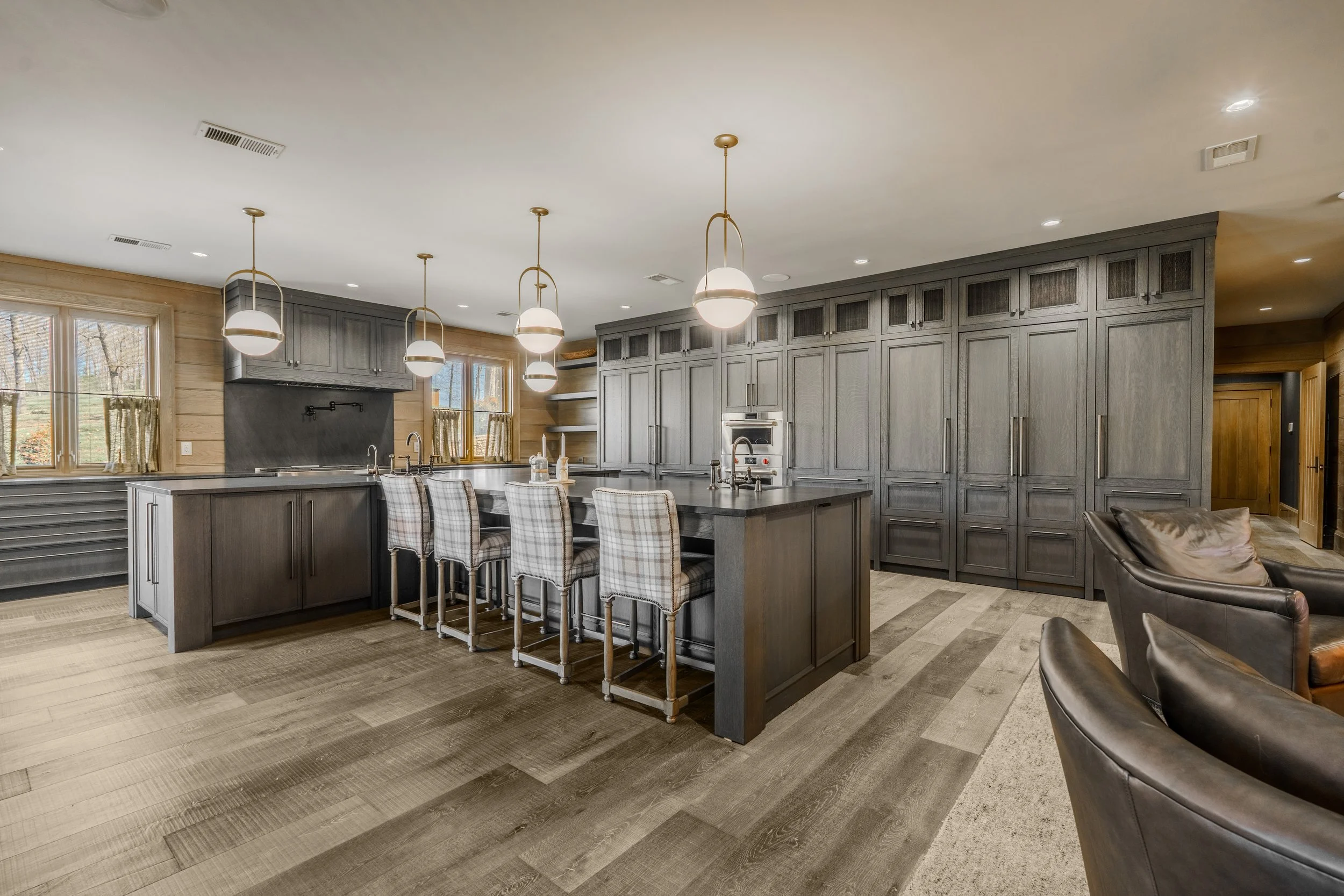Modern kitchen with gray cabinets, a large island with a dark countertop, plaid bar stools, pendant lighting, and wooden floors.