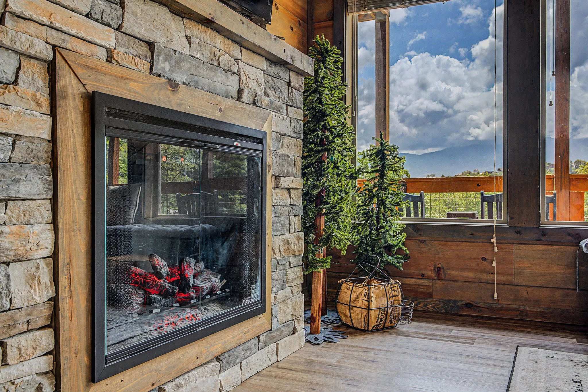 A stone and wood fireplace with glowing embers inside, on a wooden porch with a view of a partly cloudy sky and mountains in the background.