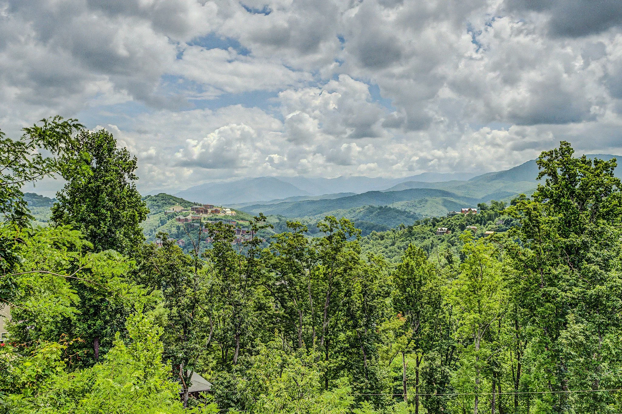 A scenic view of a lush green hilly landscape with scattered houses and trees, under a cloudy sky.