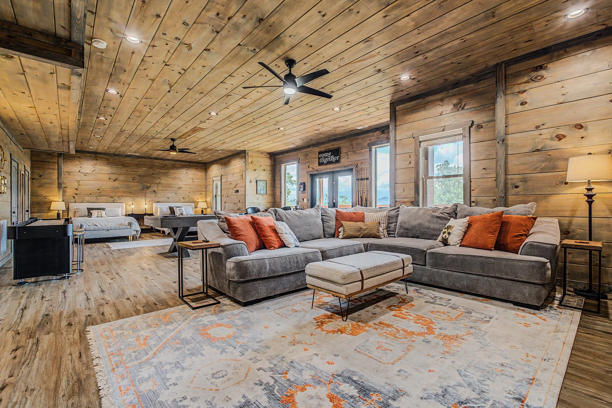 A spacious living room in a rustic style with wooden paneled ceiling and walls, featuring a large sectional sofa with orange and striped pillows, a patterned area rug, multiple windows, ceiling fans, and soft lighting.