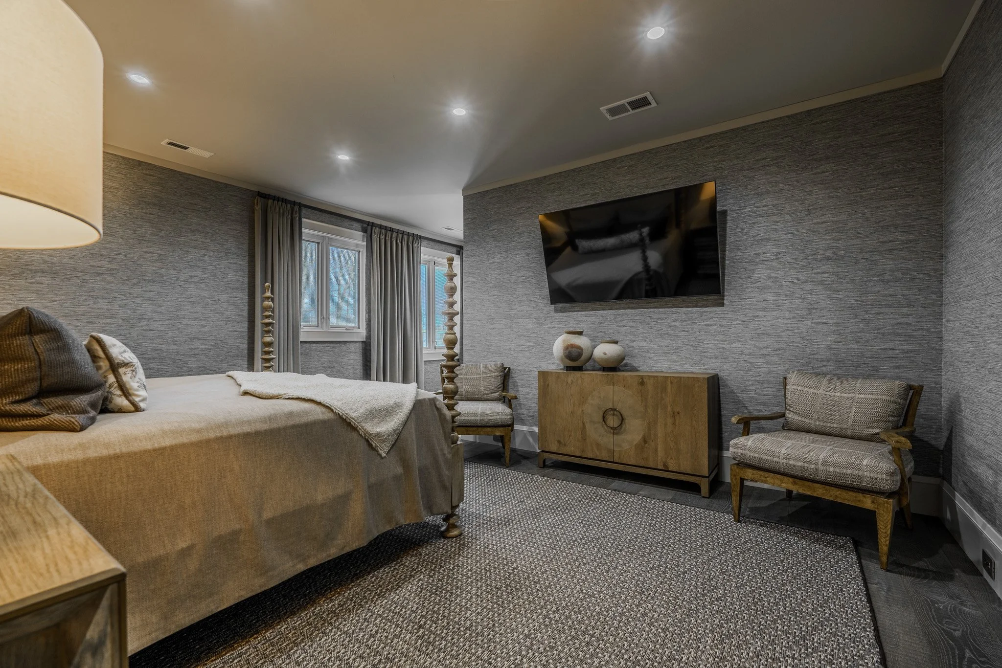 A cozy bedroom with a bed, window with curtains, a flat-screen TV on the wall, and wooden furniture, including a bench and chairs.