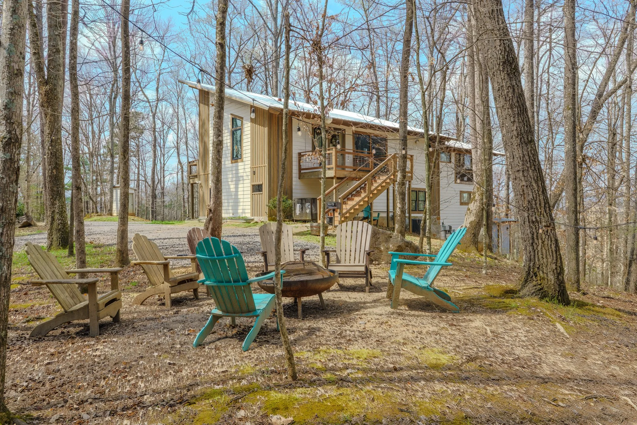 A wooded backyard with a fire pit surrounded by colorful Adirondack chairs, overlooking a modern two-story house with an outdoor staircase, set in a leafless forest under a blue sky.