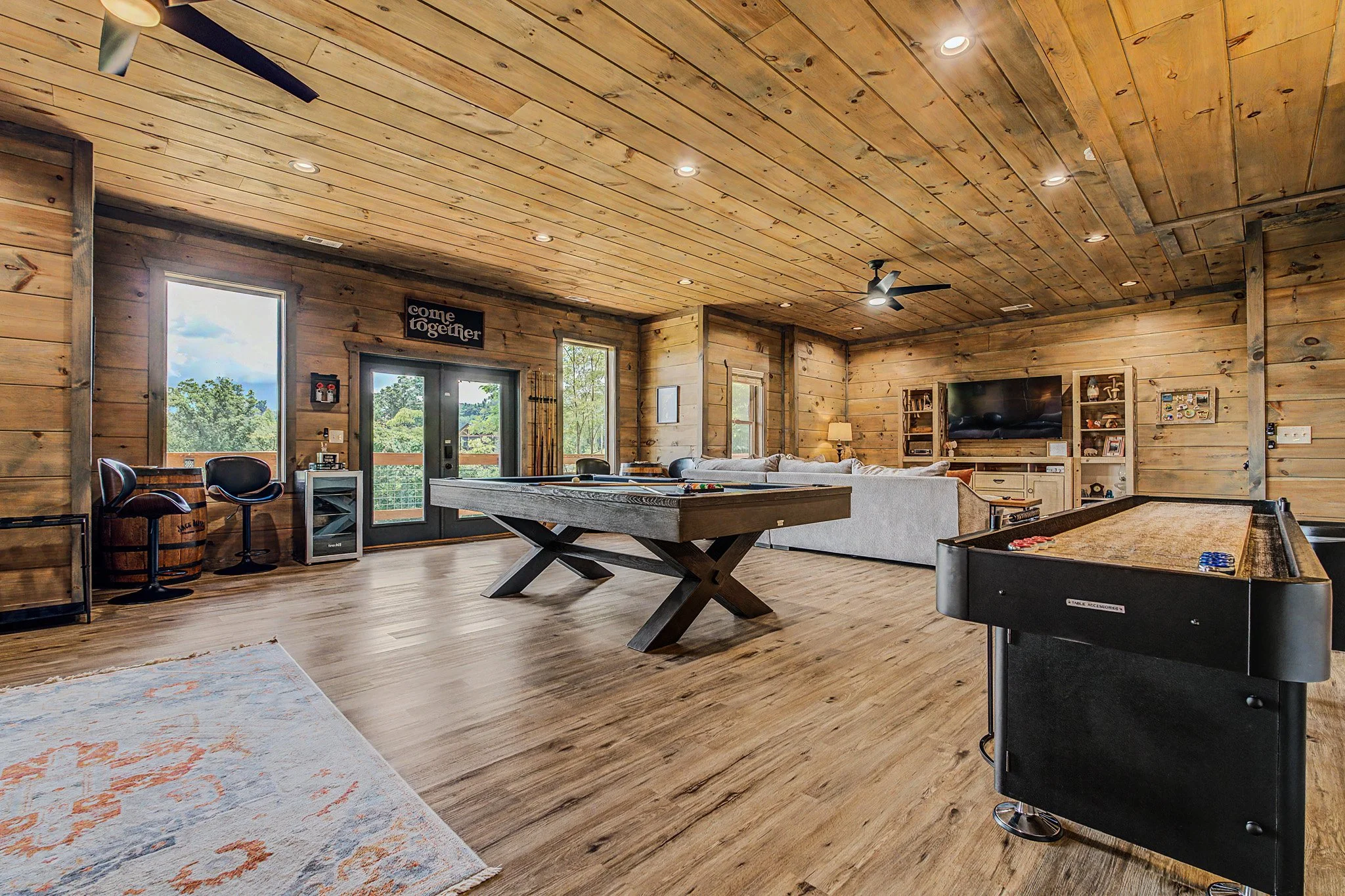 A spacious room with wooden walls and ceiling, featuring a modern ceiling fan, recessed lighting, large windows, a sitting area with a couch, a pool table, and an electronic dartboard.