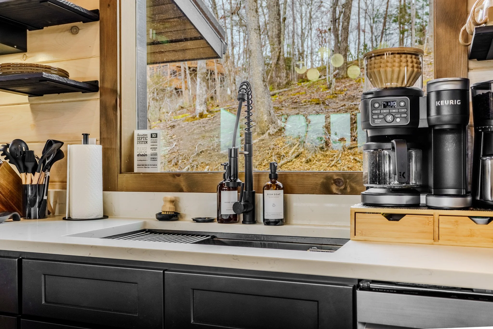 Kitchen countertop with a sink, black faucet, liquid soap dispenser, hand soap, paper towel holder, utensils, and a coffee brewing machine. A window shows a wooded outdoor scene.