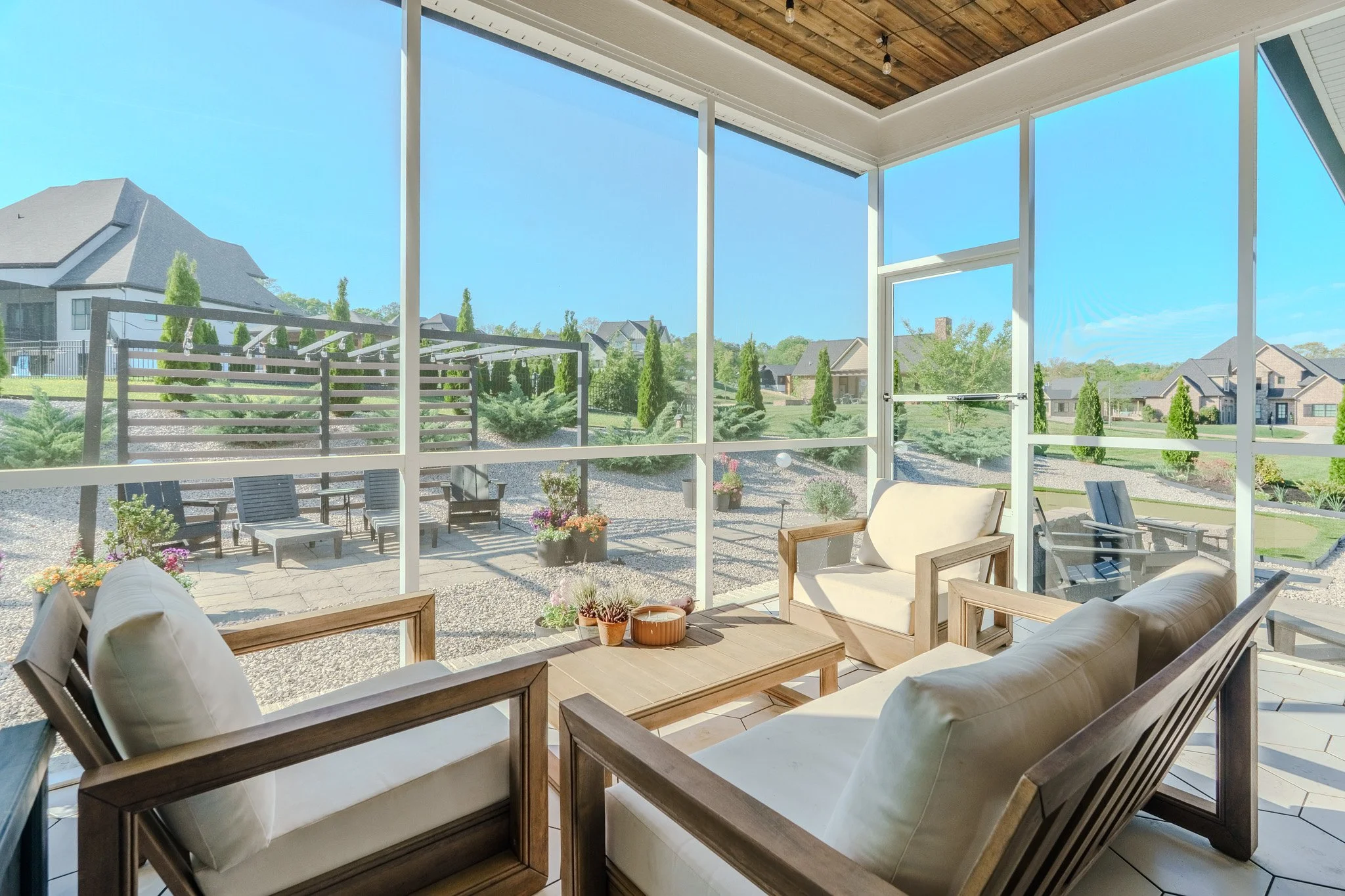 Sunroom with screened glass walls and outdoor patio furniture, including cushioned wooden chairs and potted plants, overlooking a landscaped backyard with trees and houses.