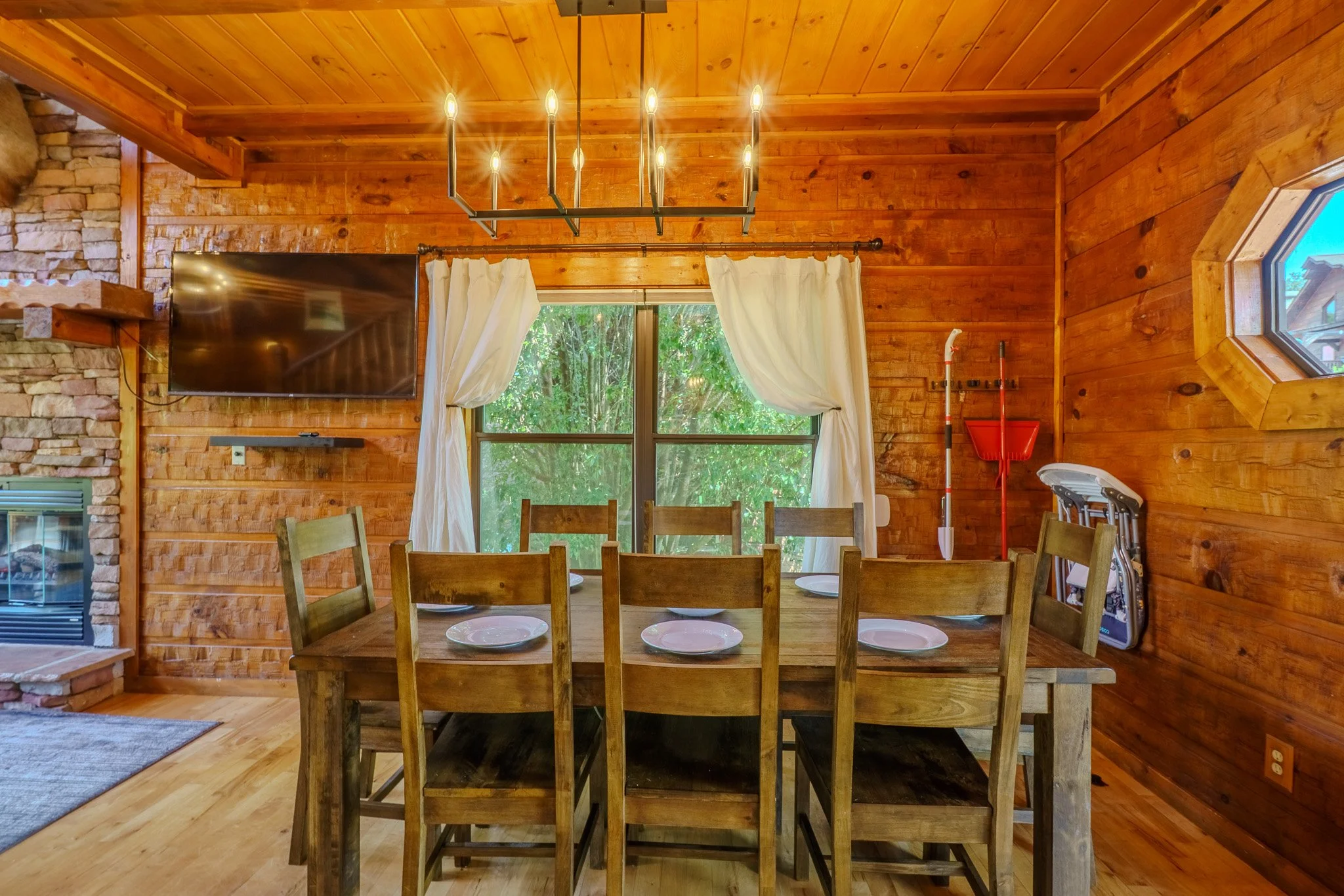 Wood-paneled dining room with six chairs around a rectangular table set with plates, decorated with a black chandelier, a window with white curtains, a wall-mounted TV, and cleaning equipment in the corner.