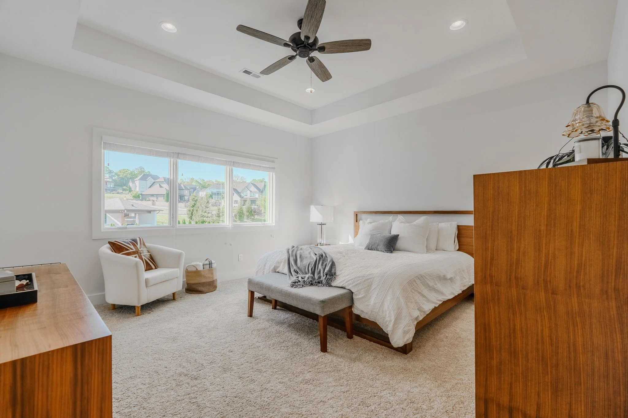 Bright bedroom with large window, white walls, ceiling fan, bed with white bedding, gray and white pillows, gray throw blanket, wooden dresser, small armchair with cushion, floor lamp, beige carpet.