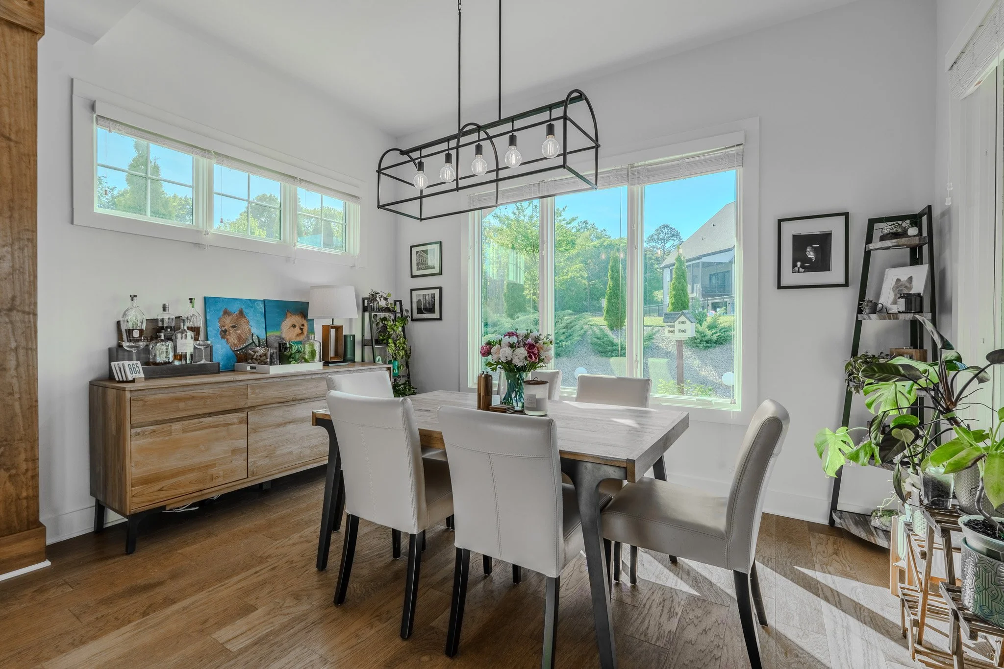 Bright dining room with a wooden table, six white chairs, a sideboard with decorative items, and large windows showing greenery outside.