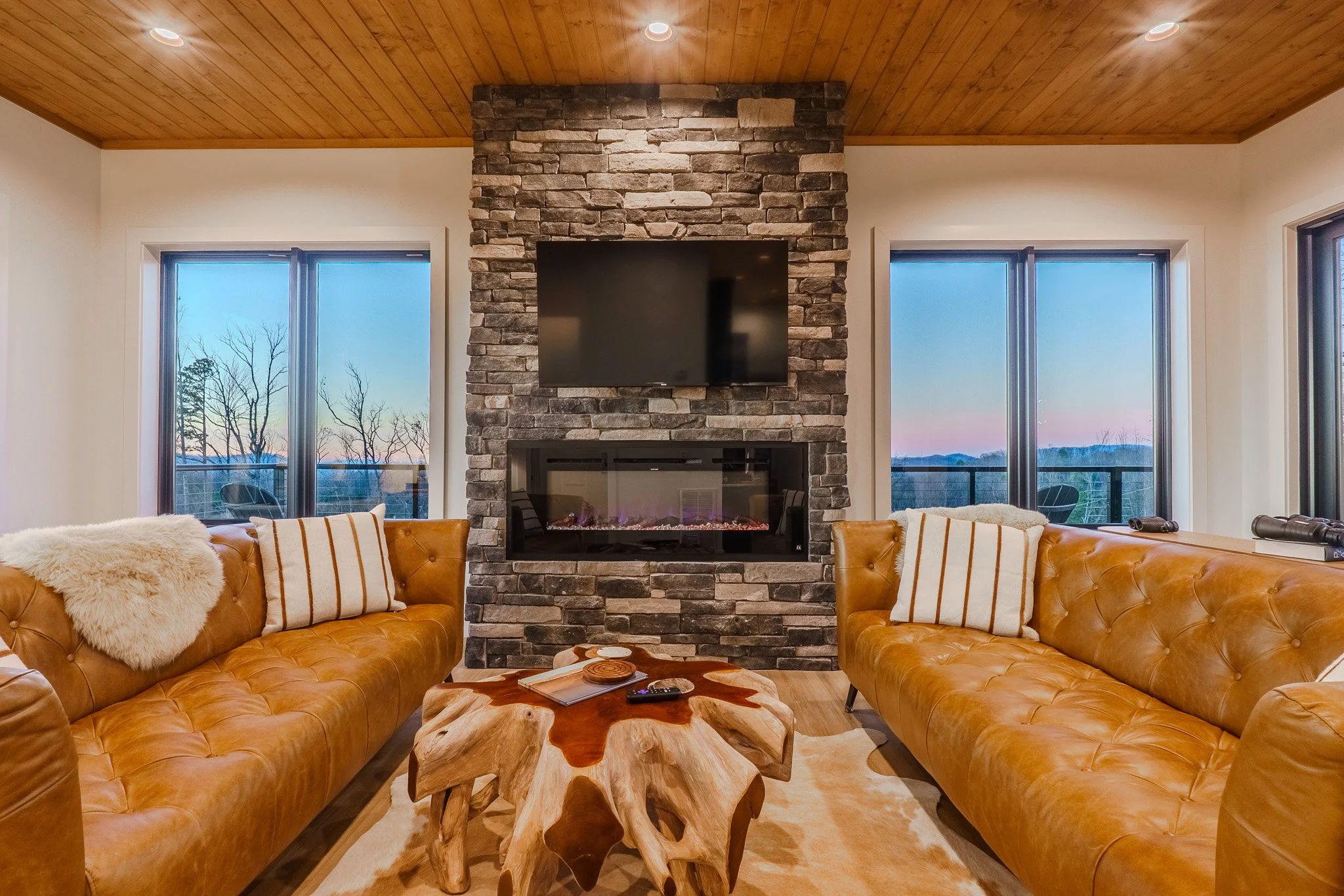 Living room with two leather sofas, a stone fireplace with a mounted TV, wooden ceiling, and large windows showing a sunset view with trees and mountains.