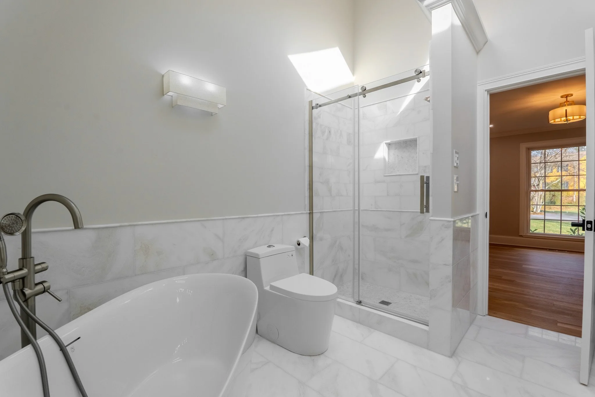 Modern bathroom with a freestanding bathtub, a toilet, and a glass-enclosed shower with marble tiles. The bathroom has white walls, a wall-mounted light, and a skylight. An open doorway reveals a room with wooden flooring and a large window with a vi