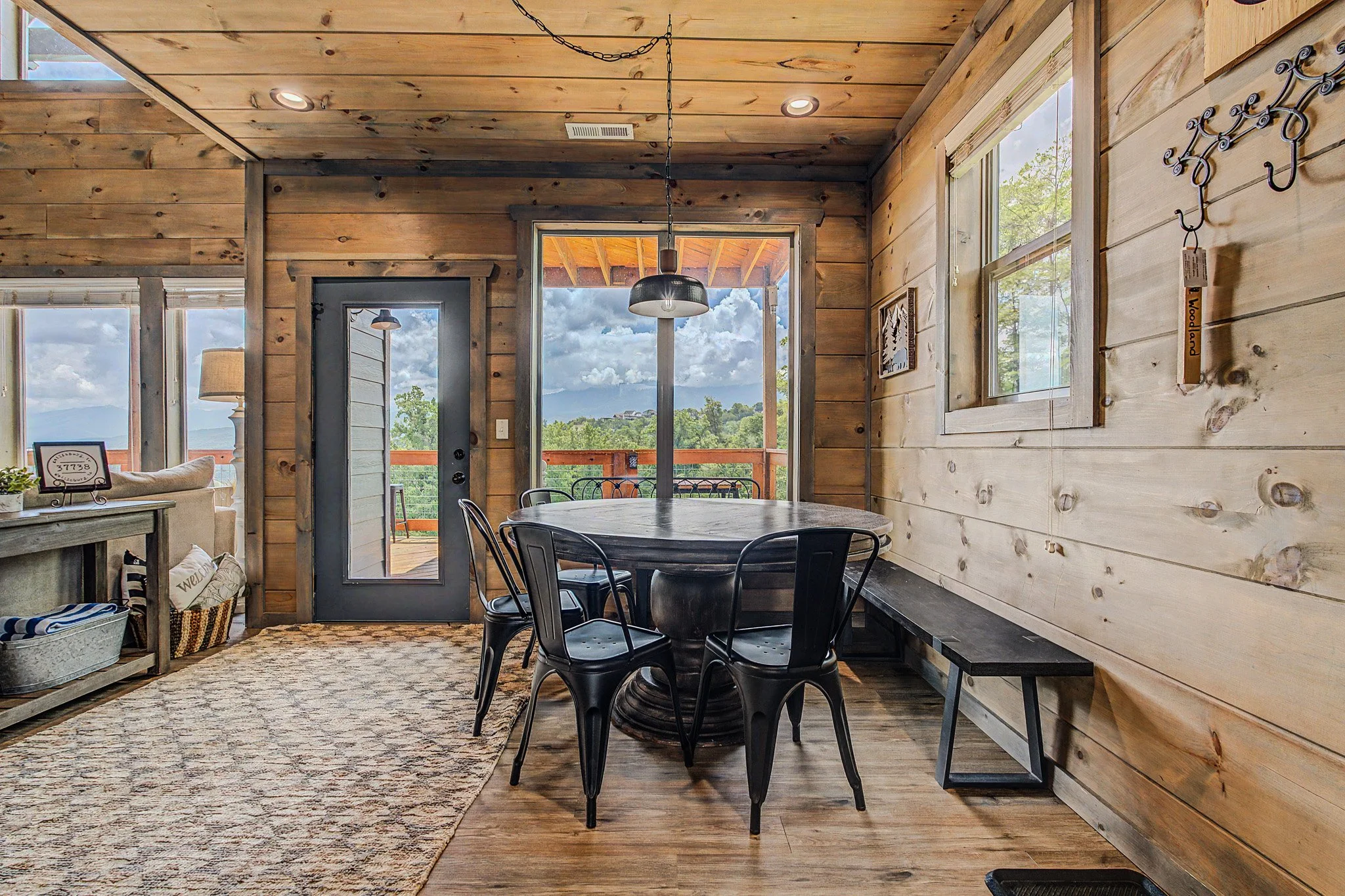 Interior of a rustic wooden cabin with a dining area, black metal chairs around a round table, large windows with scenic mountain view, and decorative hooks on the wall.