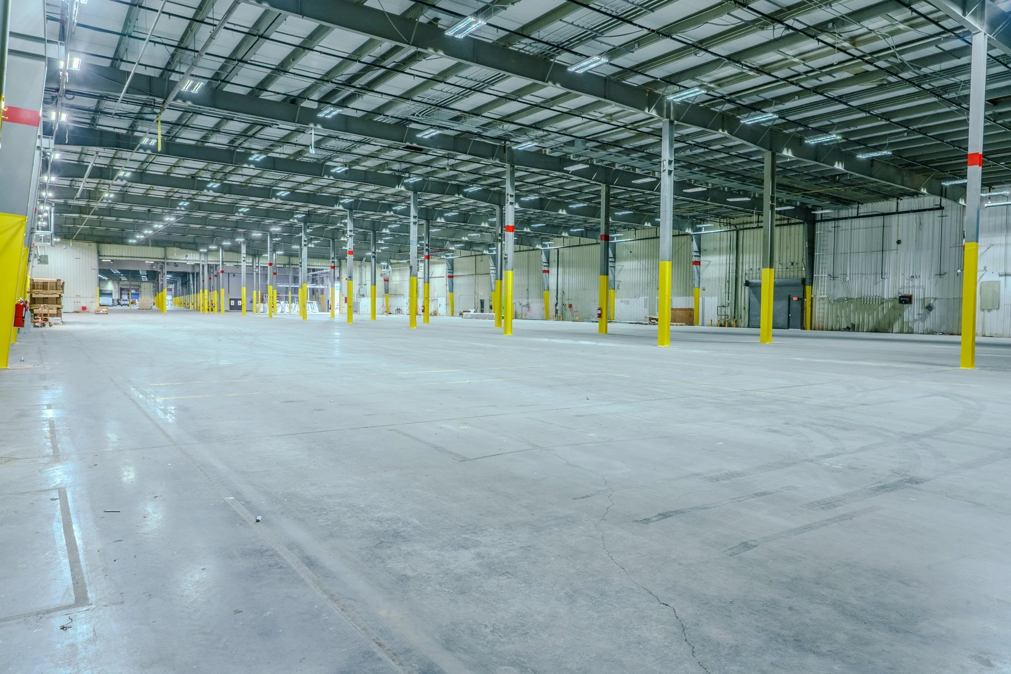 Empty indoor warehouse with concrete floor, yellow safety posts, metal support columns, and a high ceiling with industrial lighting.