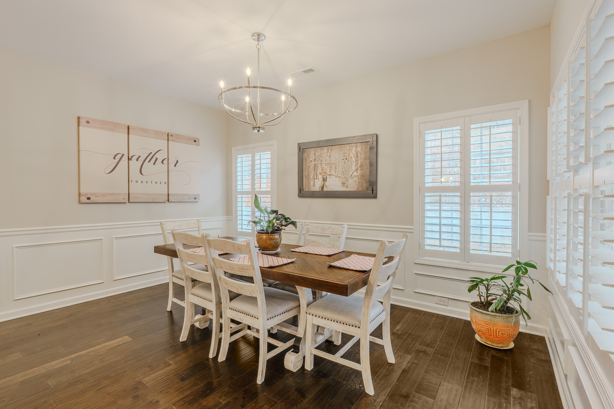 A dining room with a wooden table and six white chairs, two potted plants, framed artwork, and a modern chandelier.