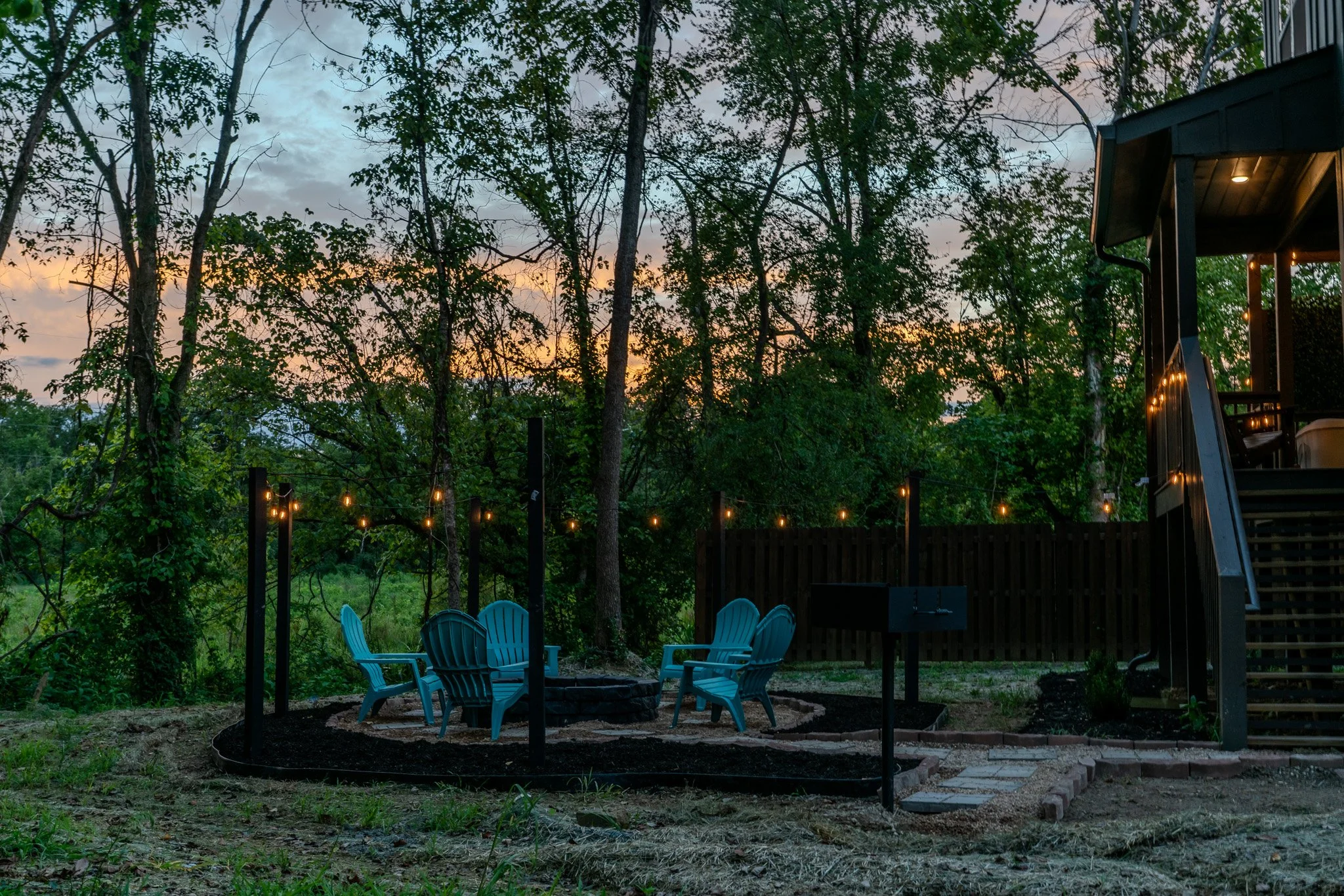Outdoor backyard seating area with five blue chairs around a fire pit, string lights hanging, trees in the background, and a deck with stairs on the right at dusk.