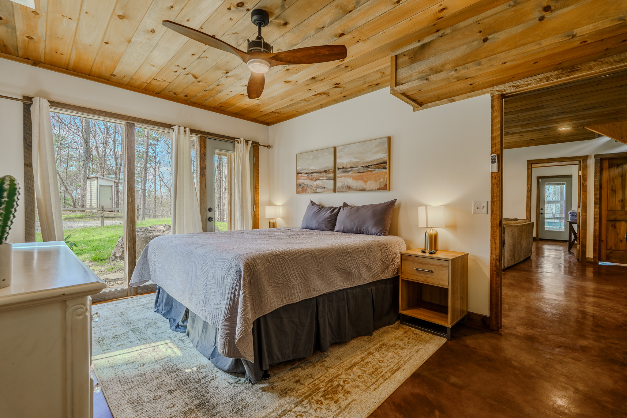 A bedroom with a wooden ceiling and floors, large windows with white curtains, a bed with lavender pillows and a beige quilt, two bedside lamps, and abstract artwork above the bed. There is a sliding door leading outside and a partially visible livin