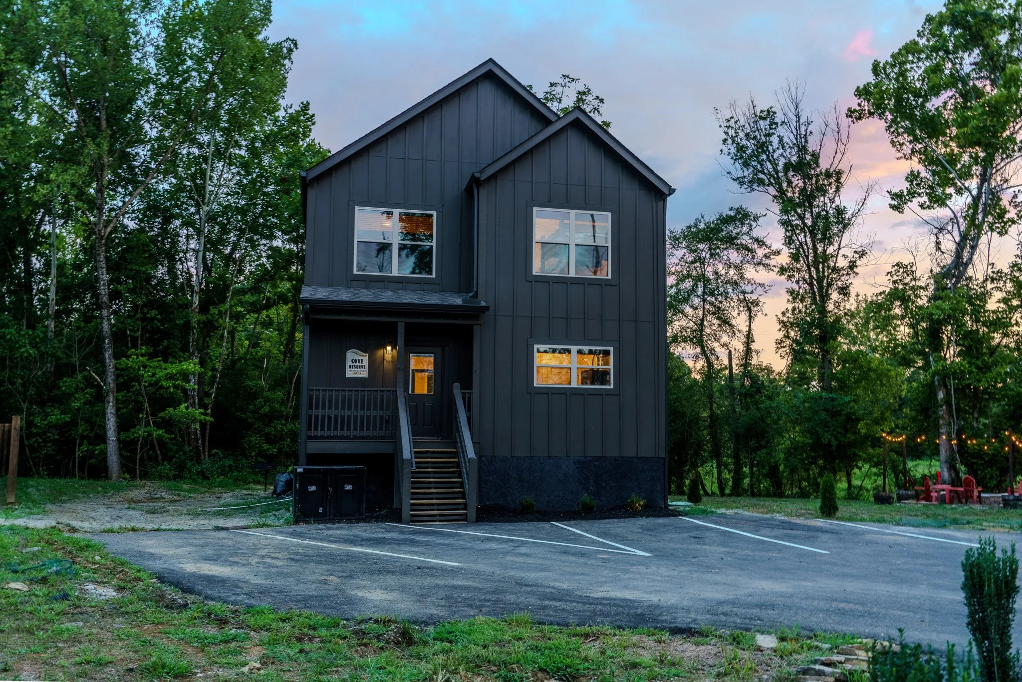 A modern black two-story house with a small front porch, surrounded by trees, with parking spaces in front and outdoor seating on the side, during dusk.