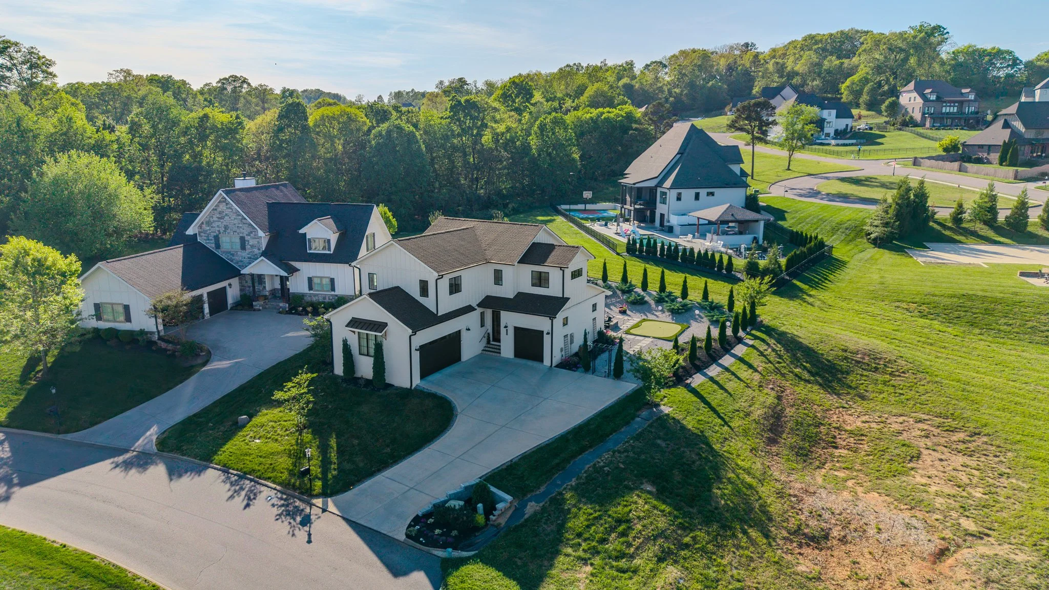 Aerial view of a large white modern house with a driveway, surrounded by greenery, with a neighboring house and a large backyard with a swimming pool and sports court.