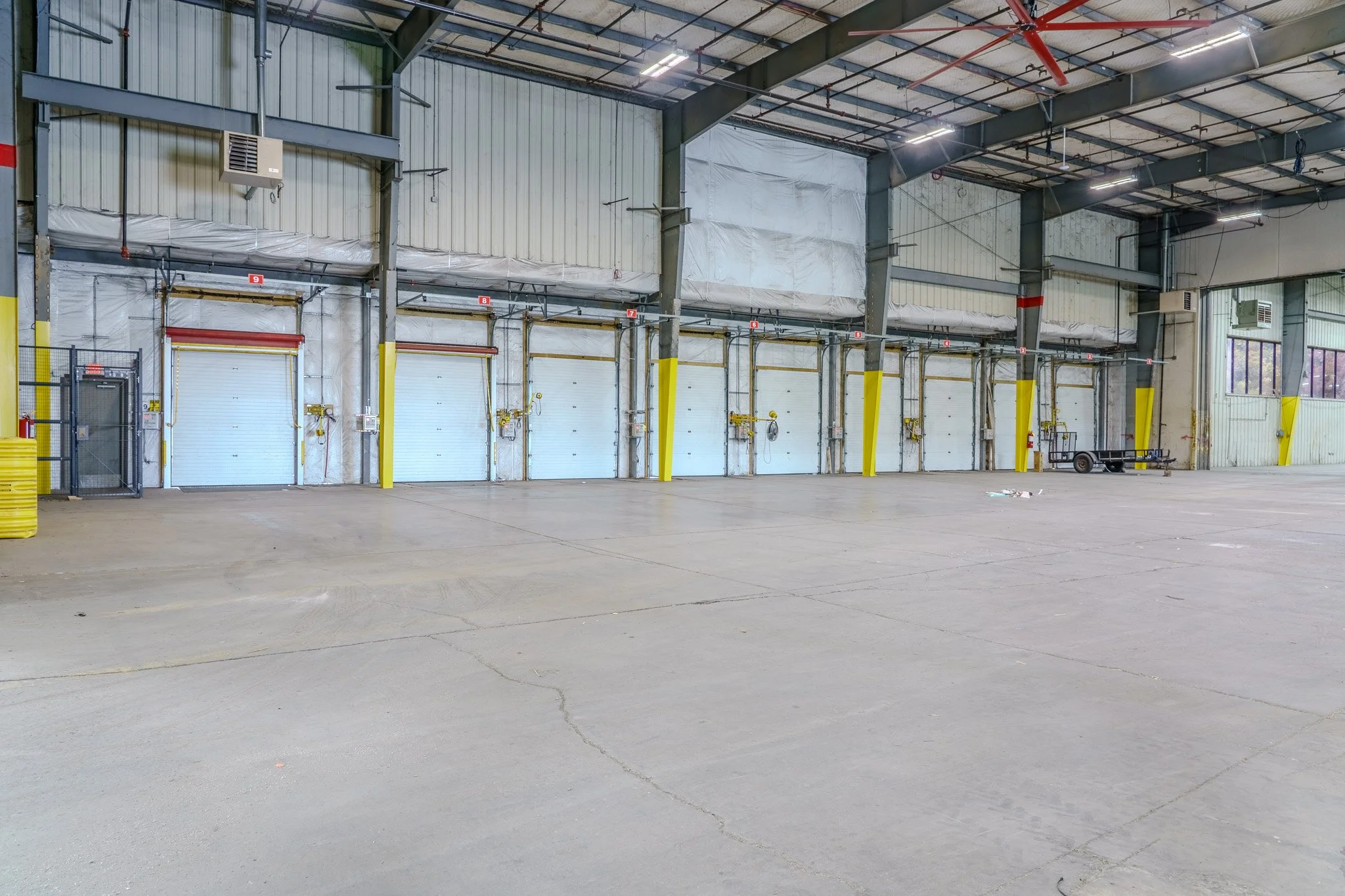 Empty warehouse with multiple loading dock doors, yellow safety barriers, and a trailer parked inside.