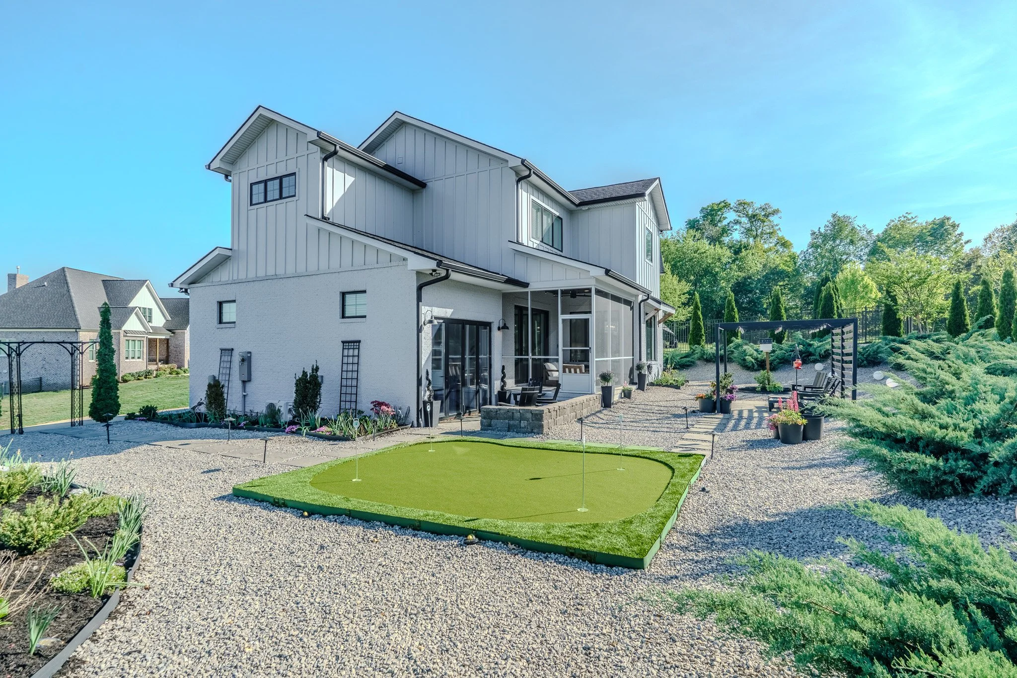 The backyard of a modern house featuring a small putting green, gravel pathways, potted plants, and outdoor seating area with a pergola, surrounded by green bushes and trees.