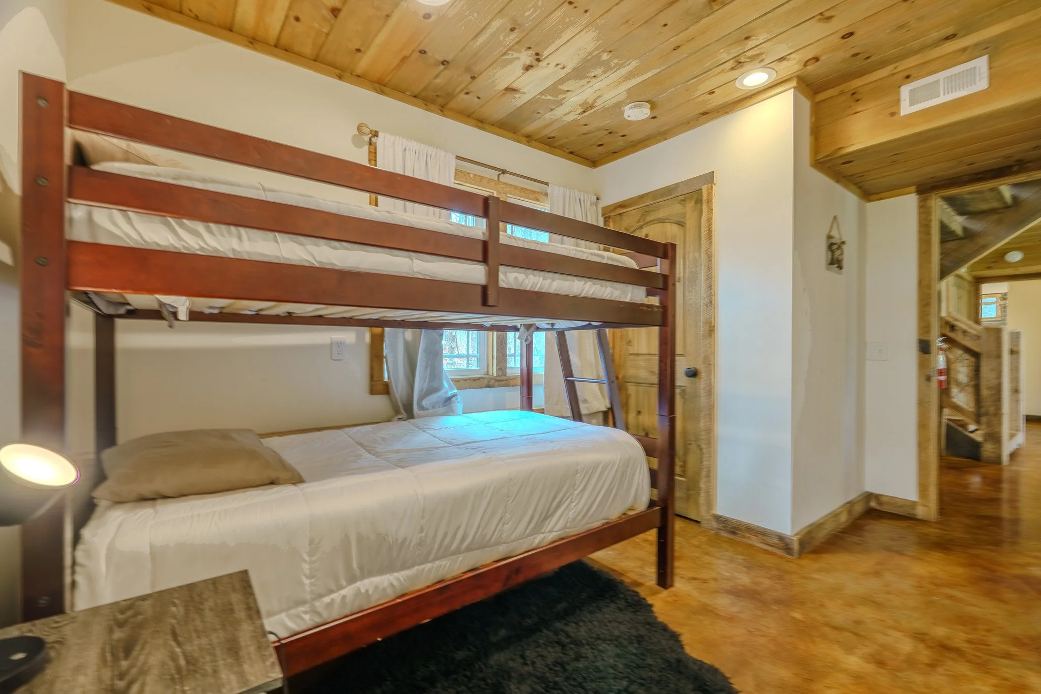 A rustic bedroom with a wooden bunk bed, a twin bed underneath, and wood paneling on the ceiling and walls. There are windows with white curtains, and a nightstand with a lamp is visible in the corner.